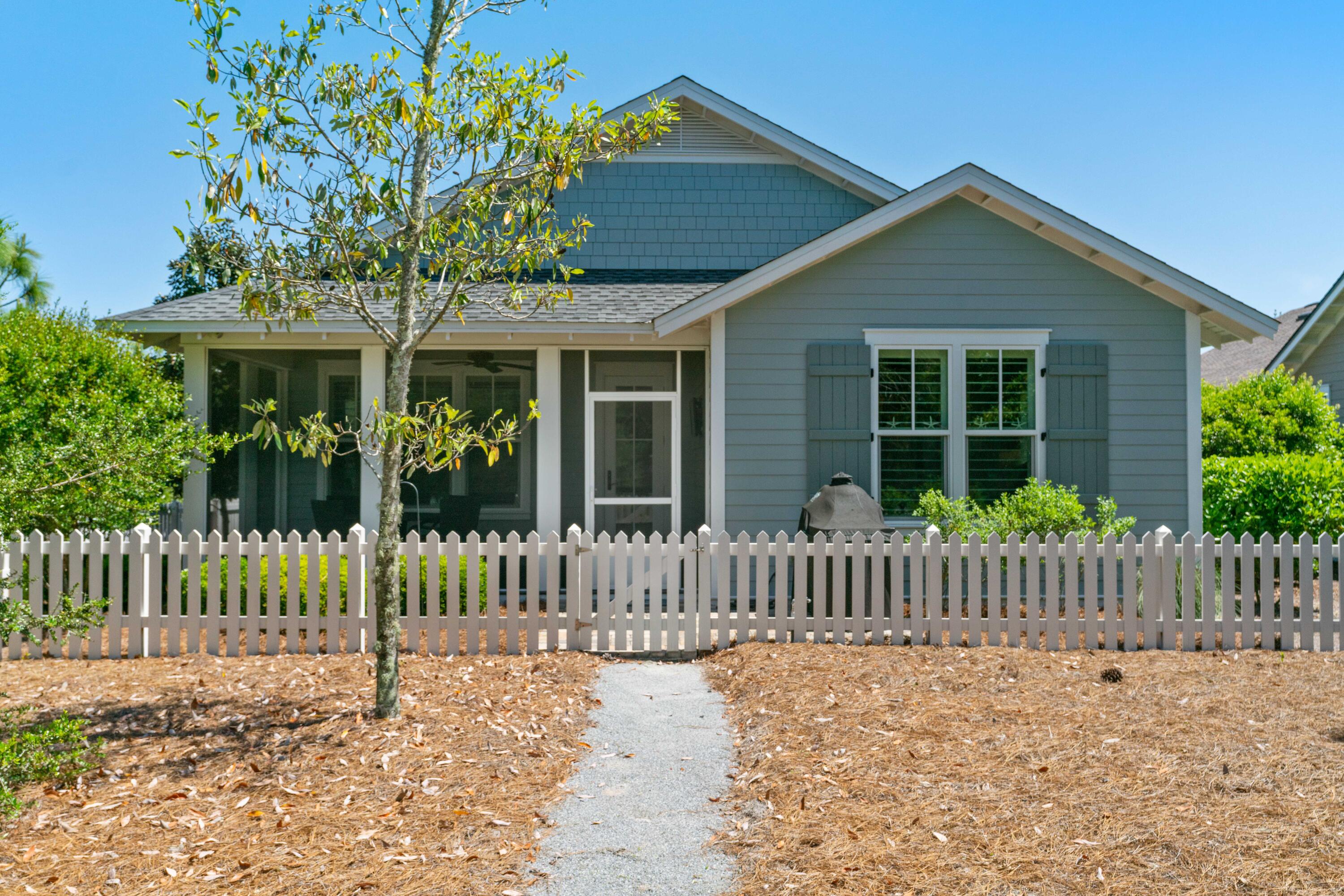 Blue-gray house with a screened porch behind a white picket fence and gravel walkway.