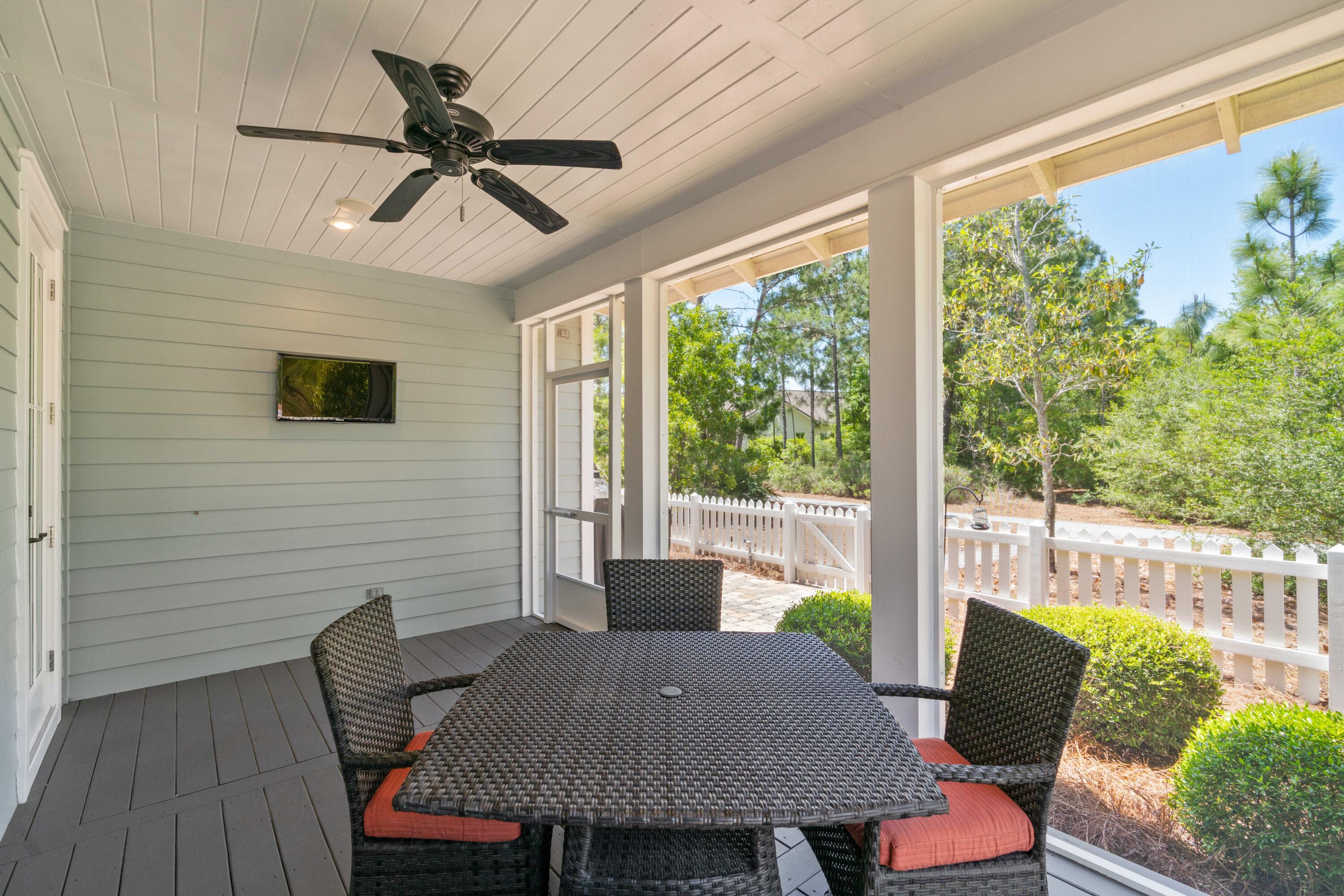 Covered patio with wicker dining set, ceiling fan, and views of trees beyond a white picket fence.
