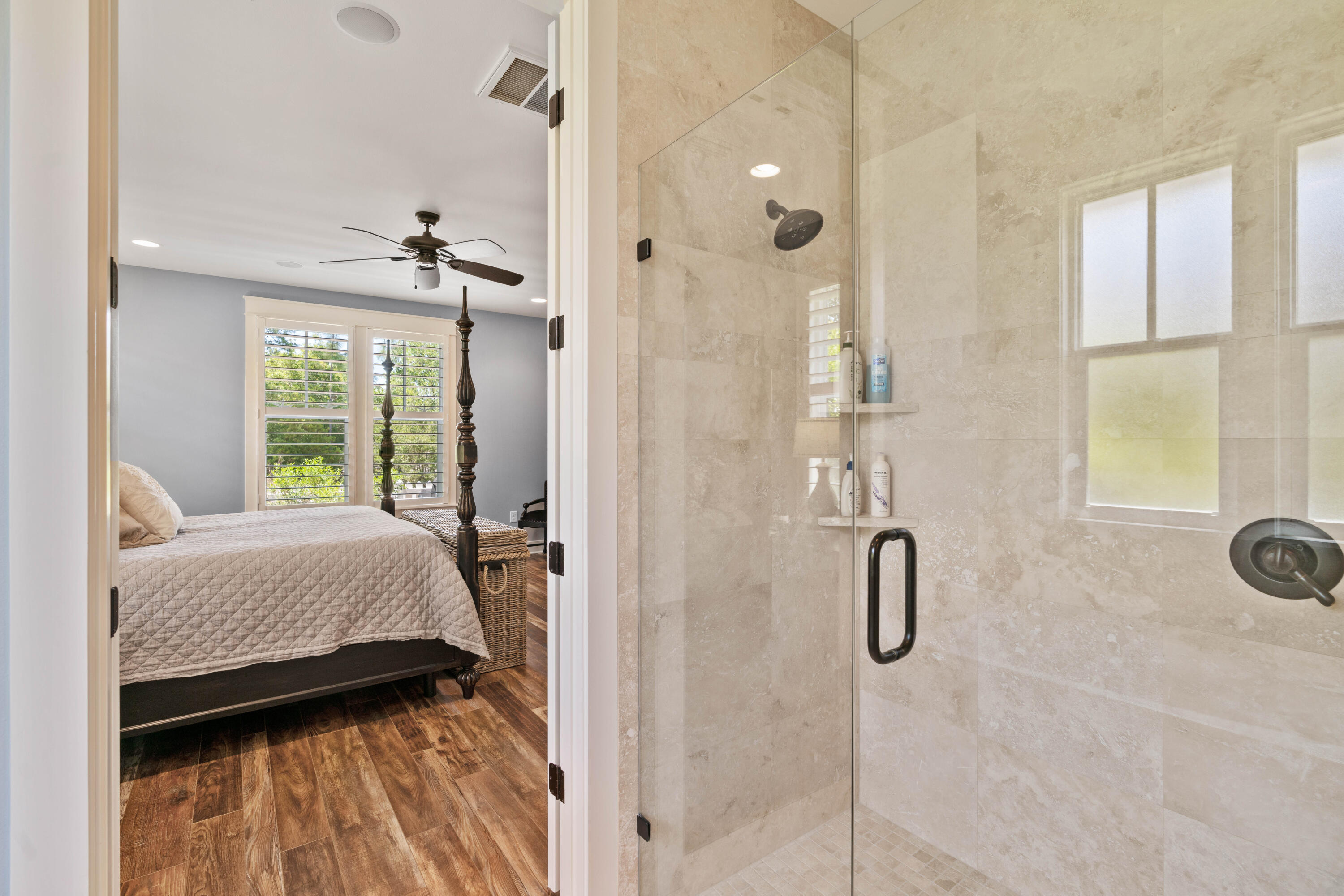 Stone-tiled glass shower beside a bedroom with a four-poster bed and plantation shutters.