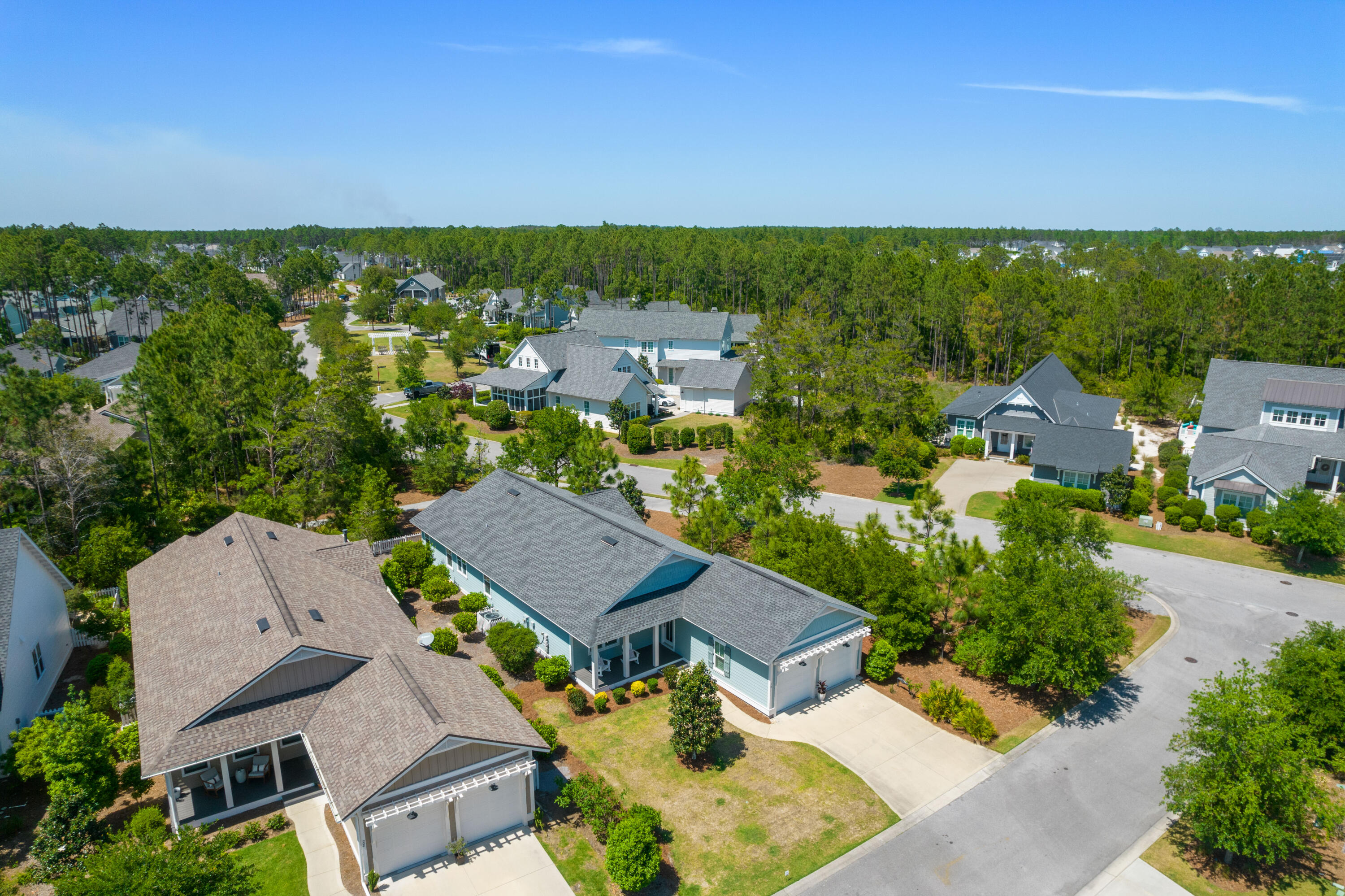 Aerial view of a blue-gray home with a covered porch, driveway, and surrounding wooded neighborhood.