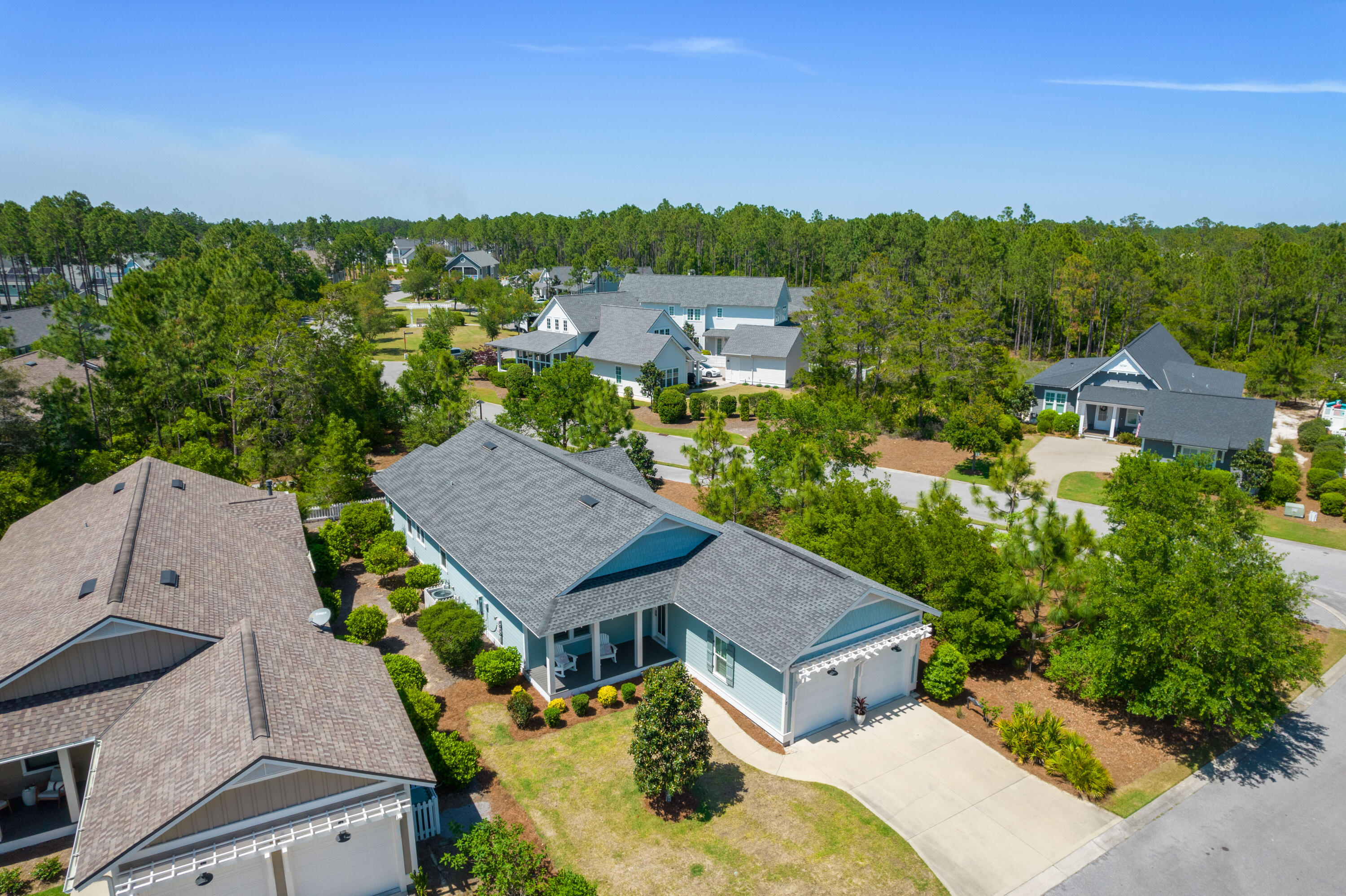 Aerial view of a blue-gray home with a front porch, garage, and curved driveway among pine trees.