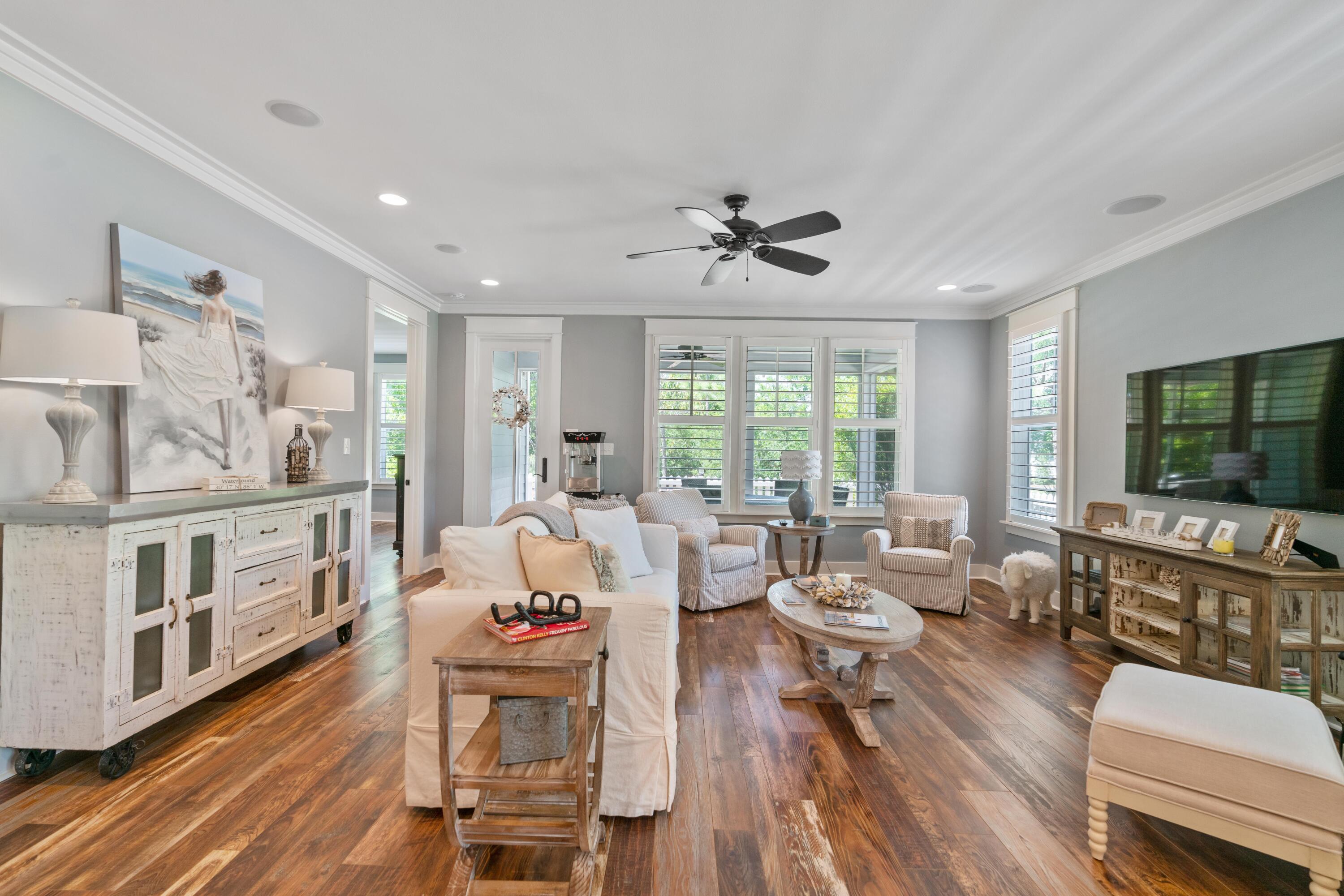 Furnished living room with shuttered windows, wood floors, cream seating, and a large television.