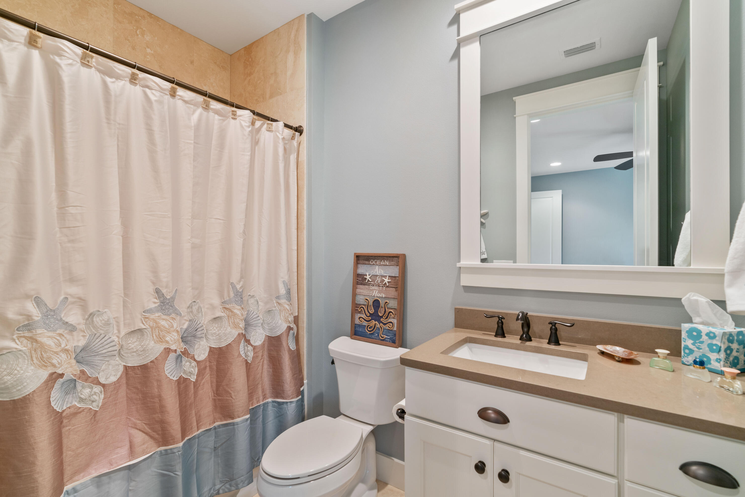 Bathroom with seashell-patterned shower curtain, white vanity, and framed coastal sign.