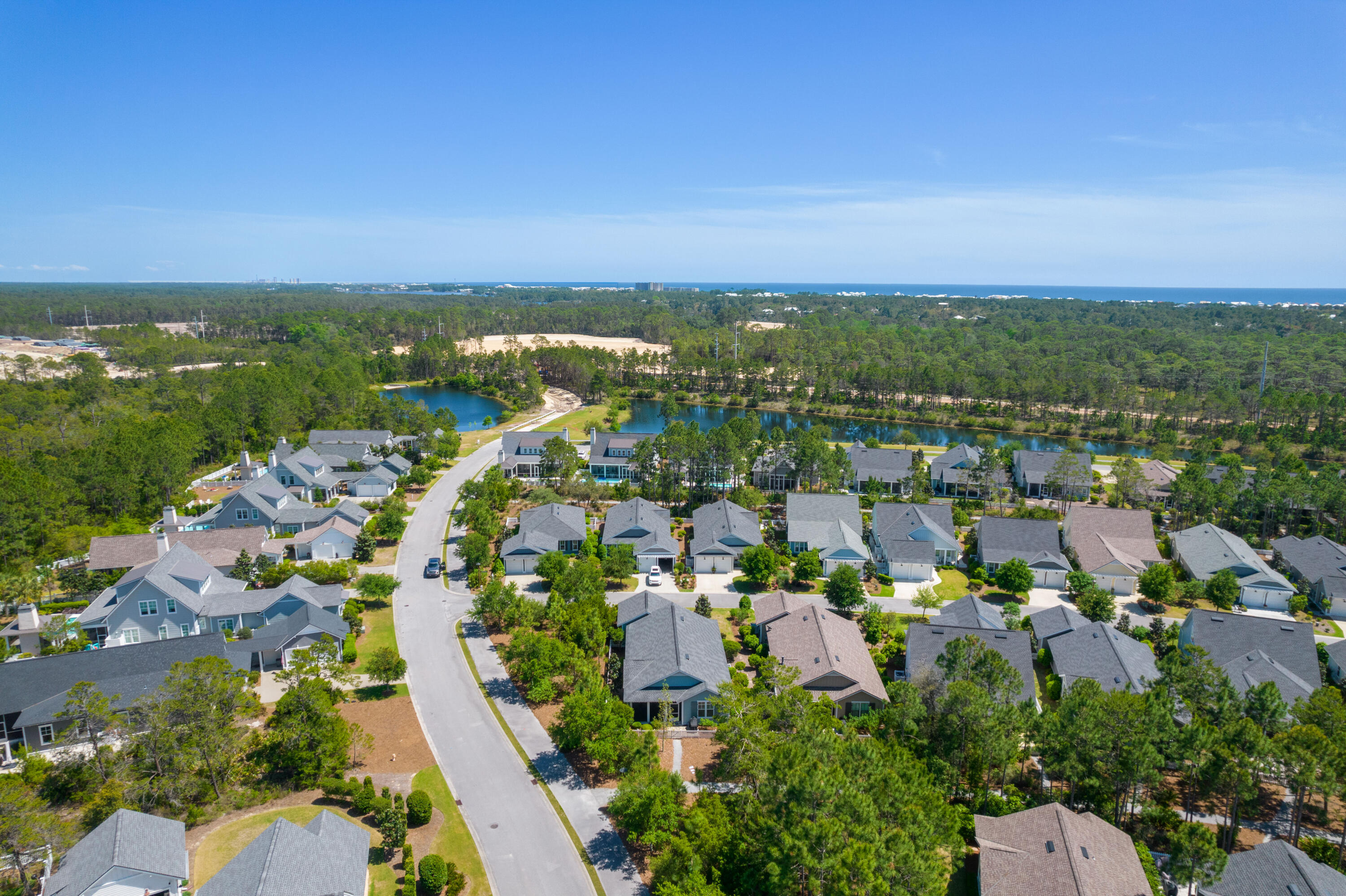 Aerial view of a coastal neighborhood with a winding road, wooded waterway, and ocean on the horizon.