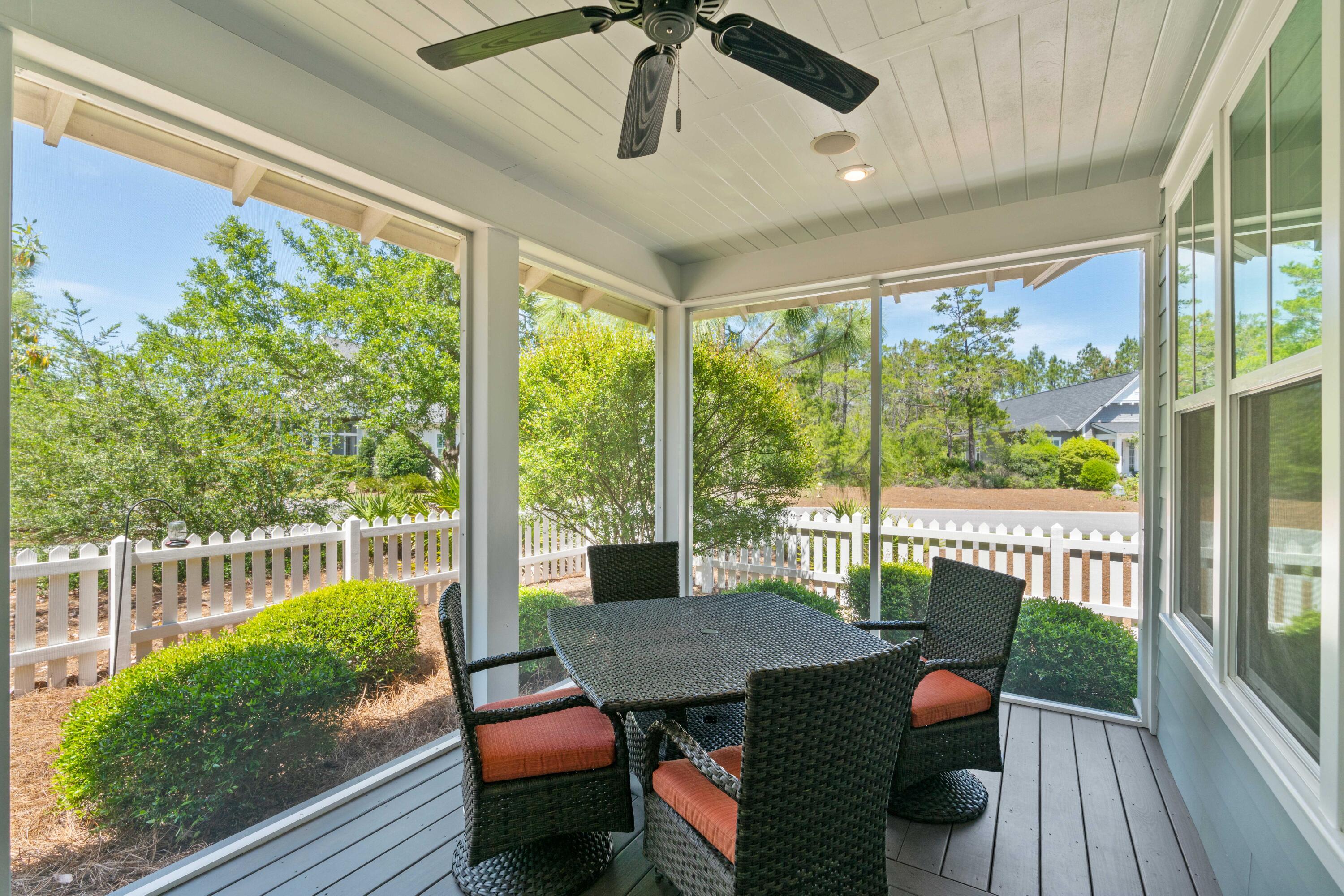 Screened porch with woven dining furniture, a ceiling fan, and views of a white picket fence and trees.