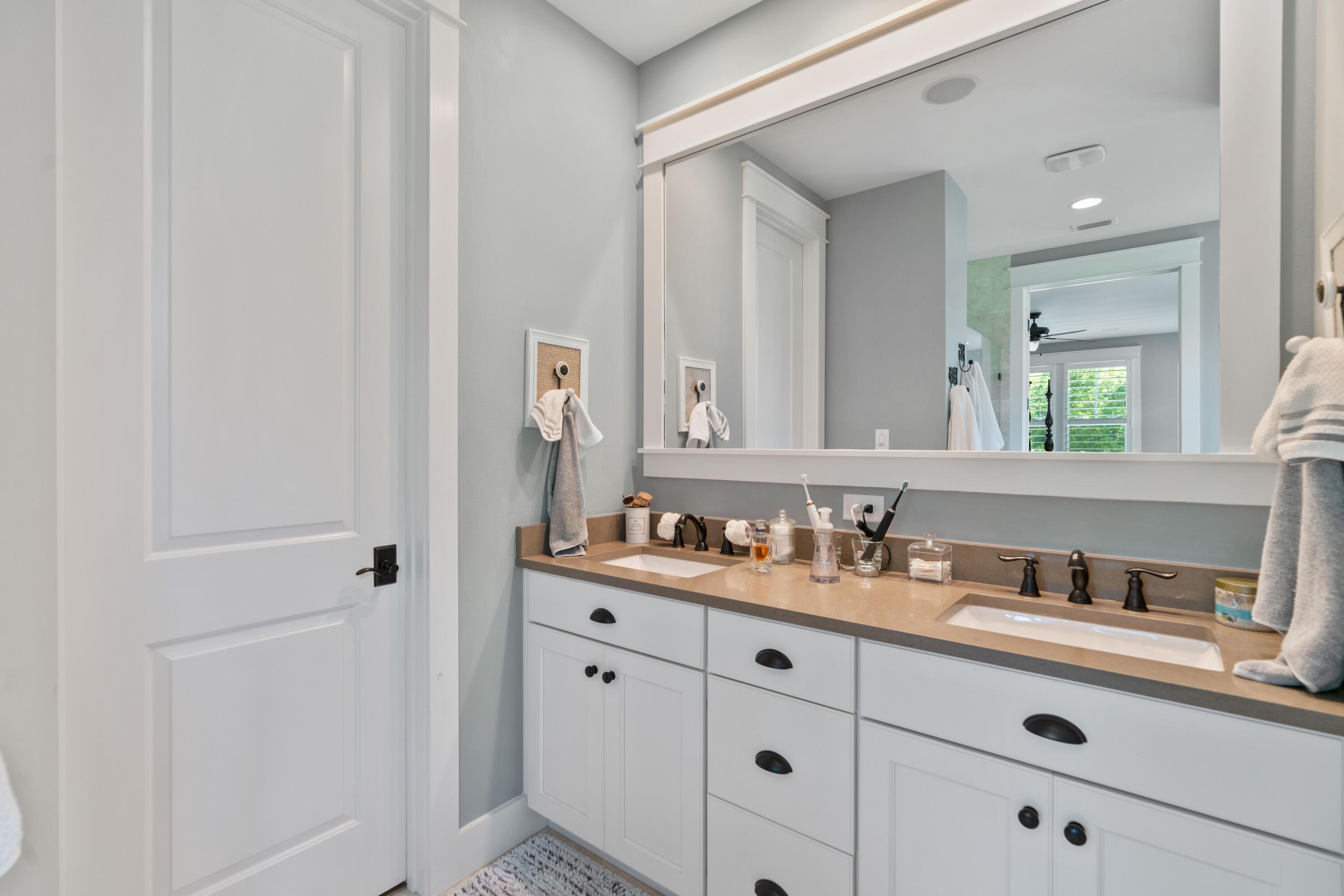 Bathroom with a long double-sink vanity, tan countertop, dark fixtures, and wide mirror.