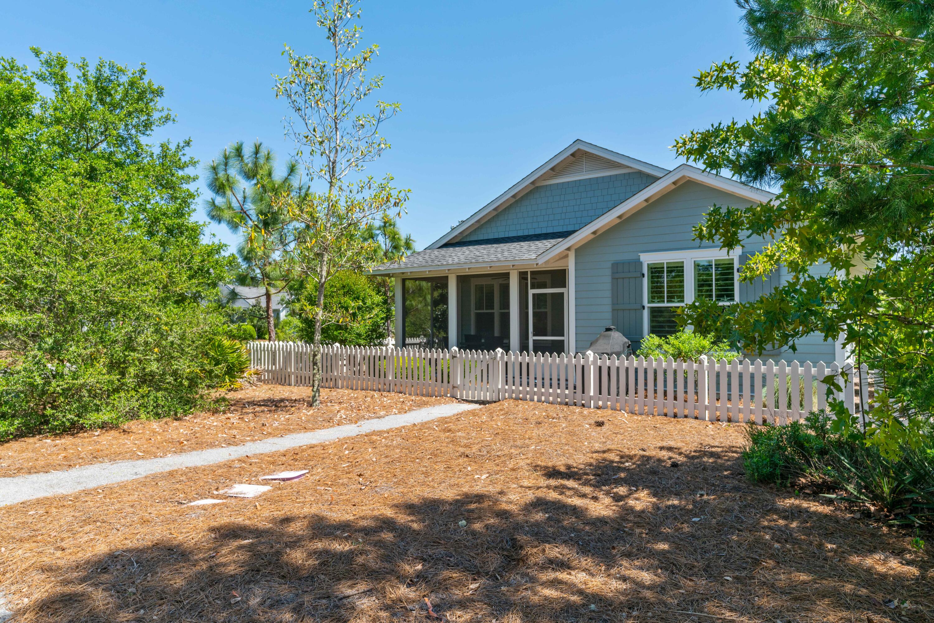 Blue-gray house with a screened porch behind a white picket fence and pine-straw yard.