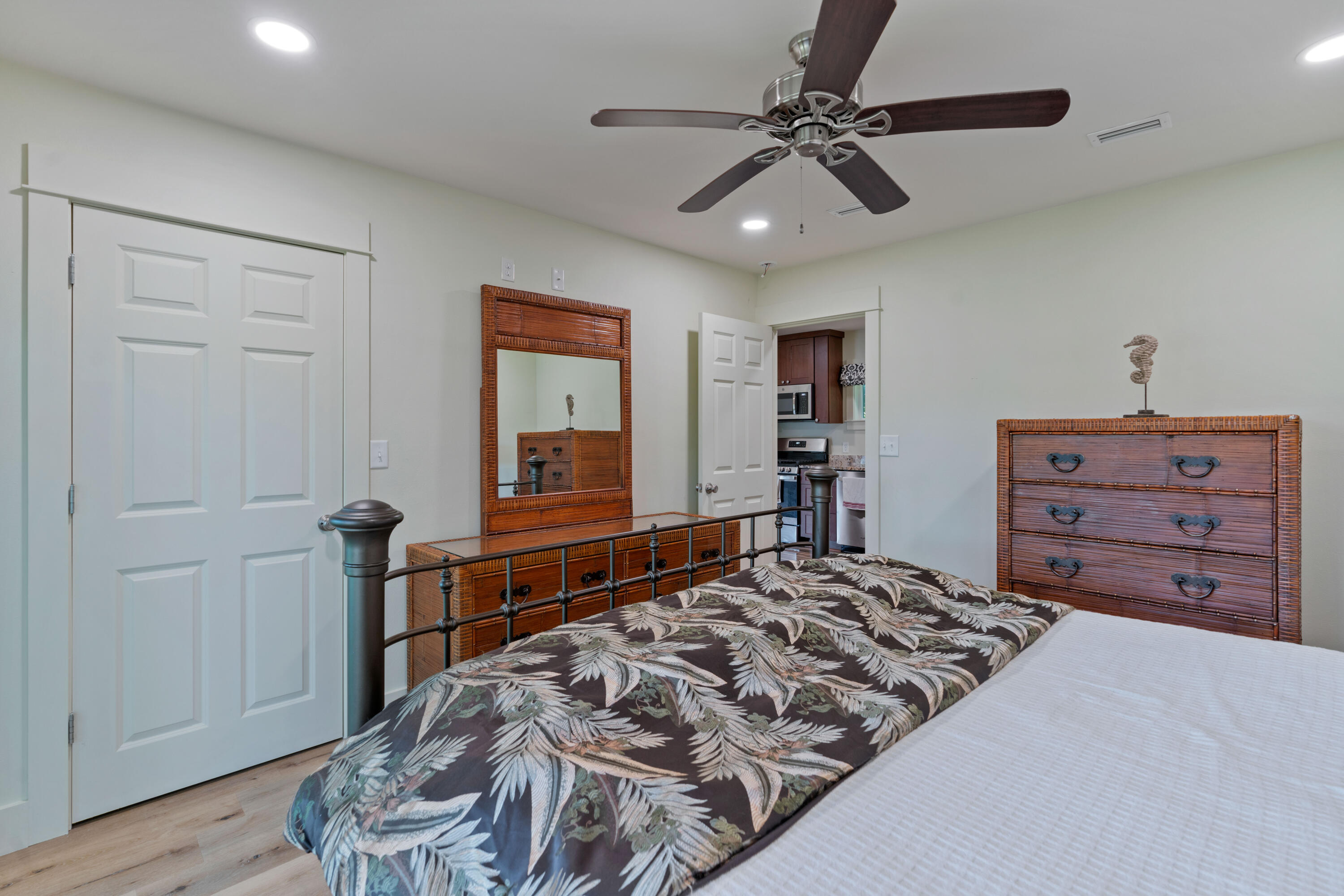 Furnished bedroom with tropical leaf bedding, woven wood dressers, and an open doorway to the kitchen.