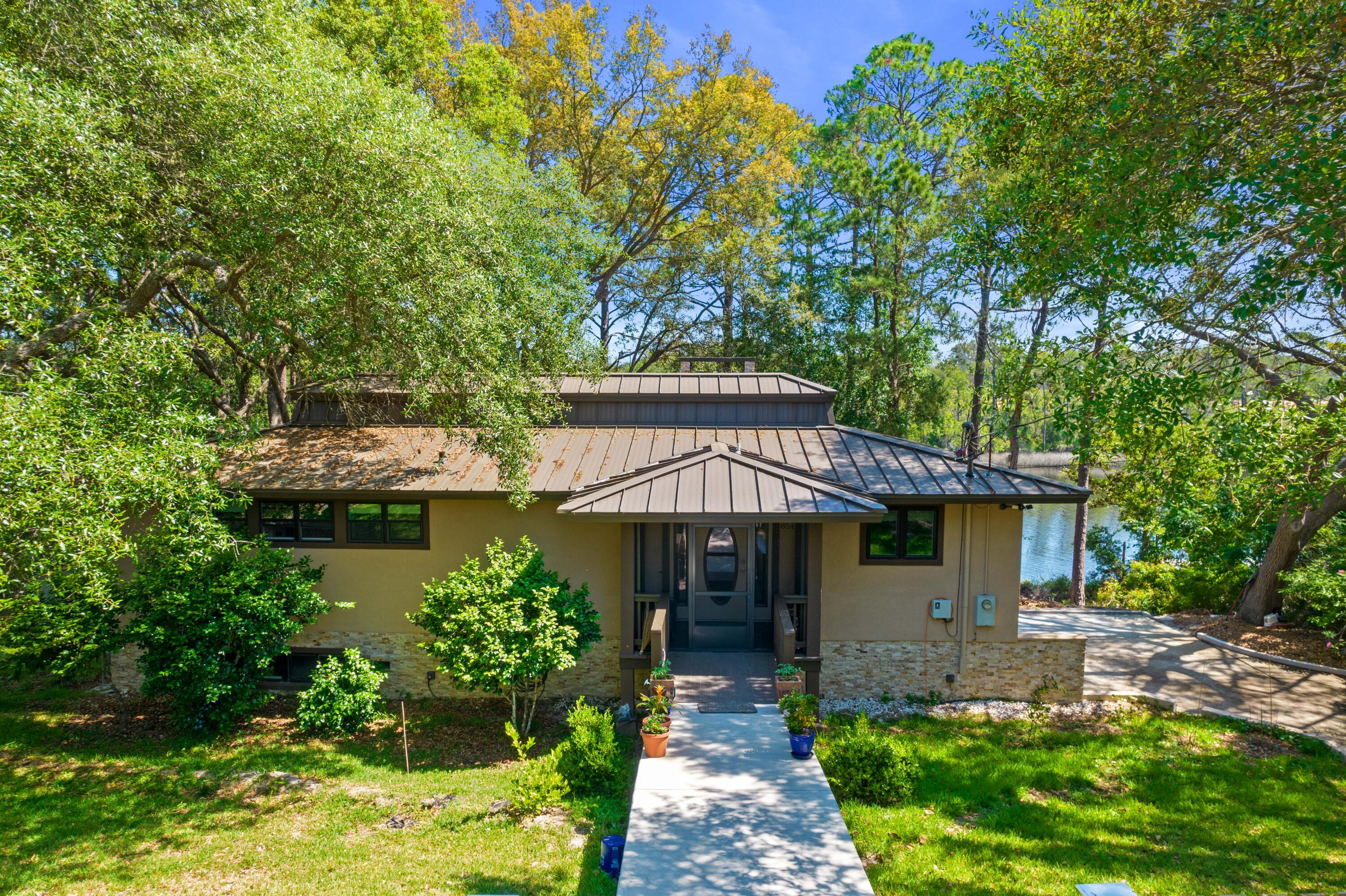 Single-story home with a metal roof, shaded front walkway, and water visible through the trees.