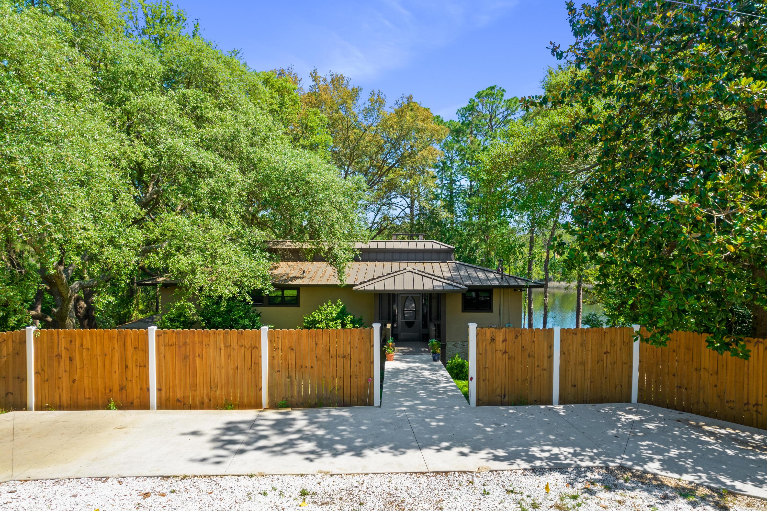 Single-story home behind a wooden fence, with a walkway to the entrance and water visible through the trees.