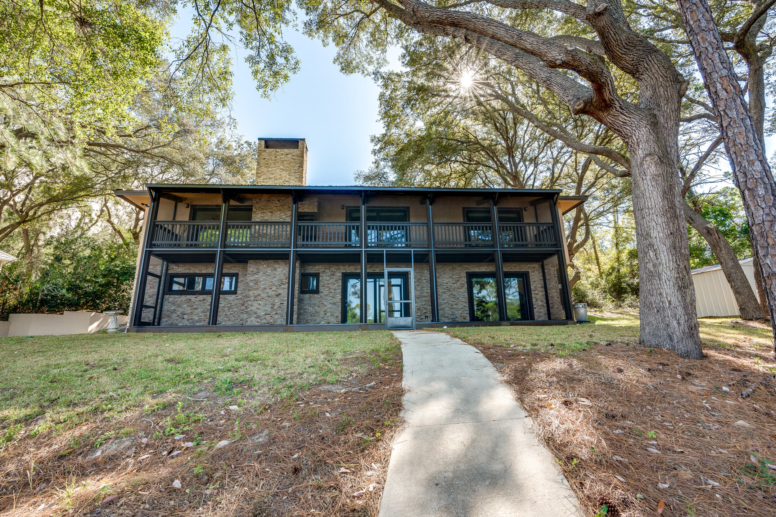 Two-story stone-and-wood home with a covered balcony and tall stone chimney beneath mature trees.