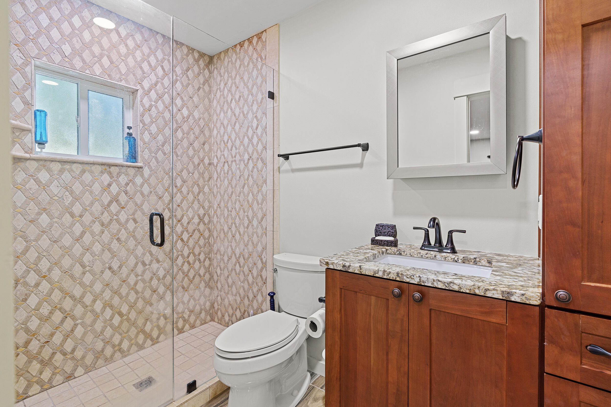 Bathroom with a patterned tile shower, white toilet, and wood vanity with granite countertop.