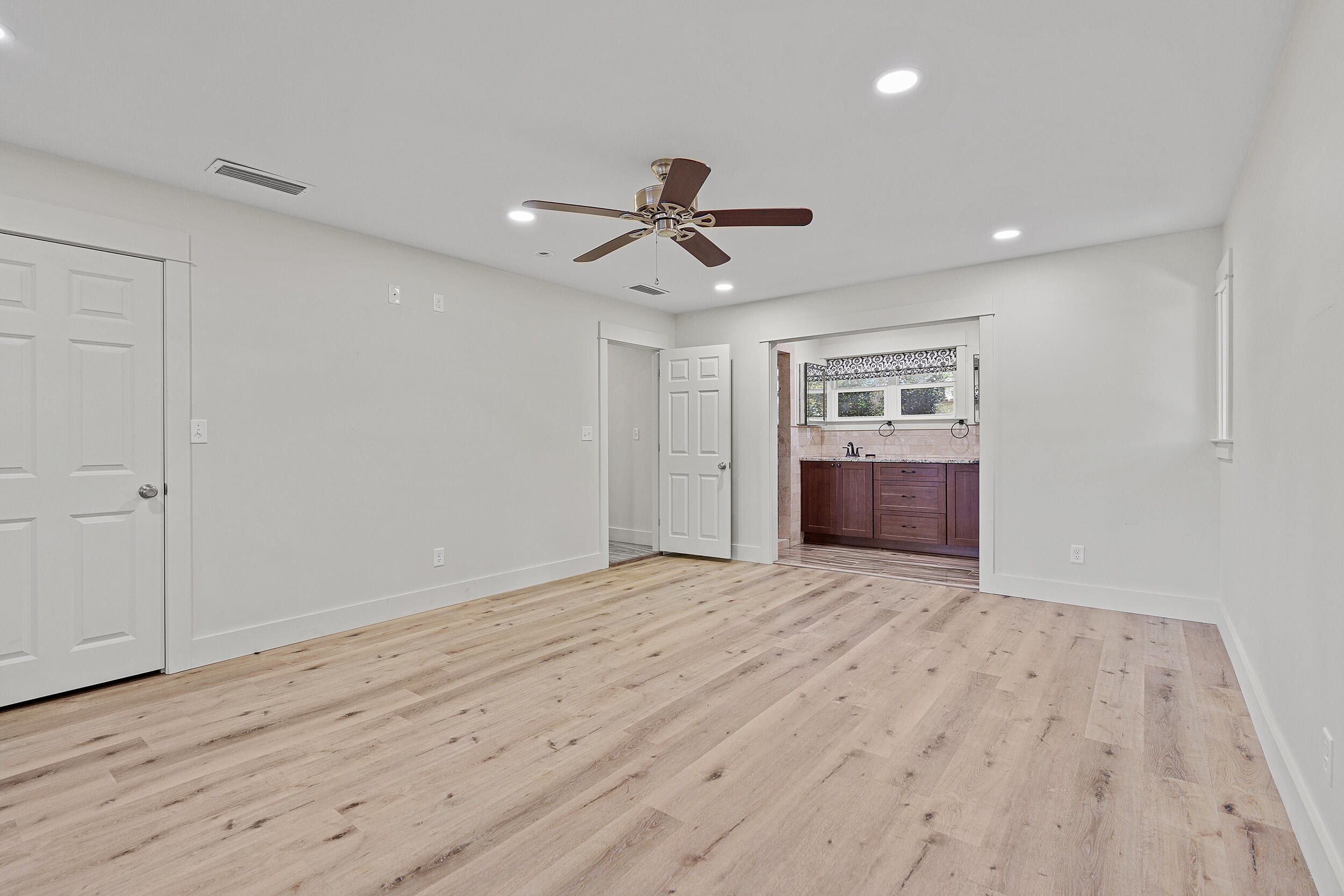 Vacant bedroom with light wood floors and ceiling fan, opening into a bathroom with dark wood cabinets.