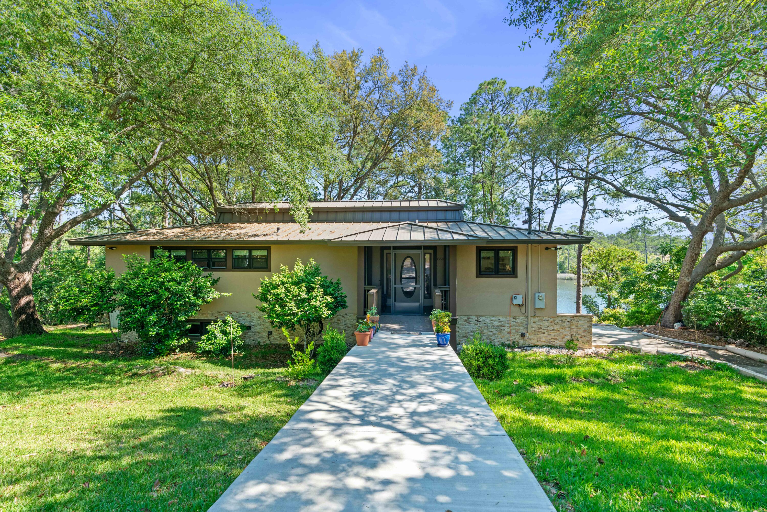 Single-story home with a metal roof and stone veneer, surrounded by trees with water visible beside it.