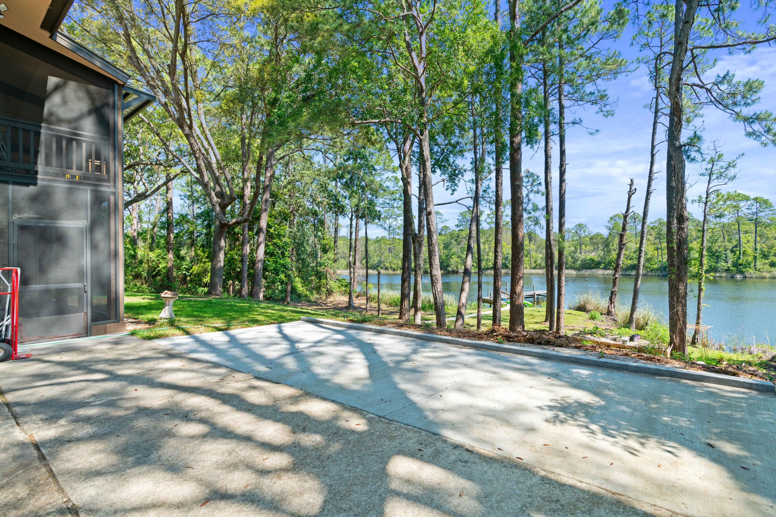 Concrete patio and driveway beside a wooded waterfront, with a small dock along the water.