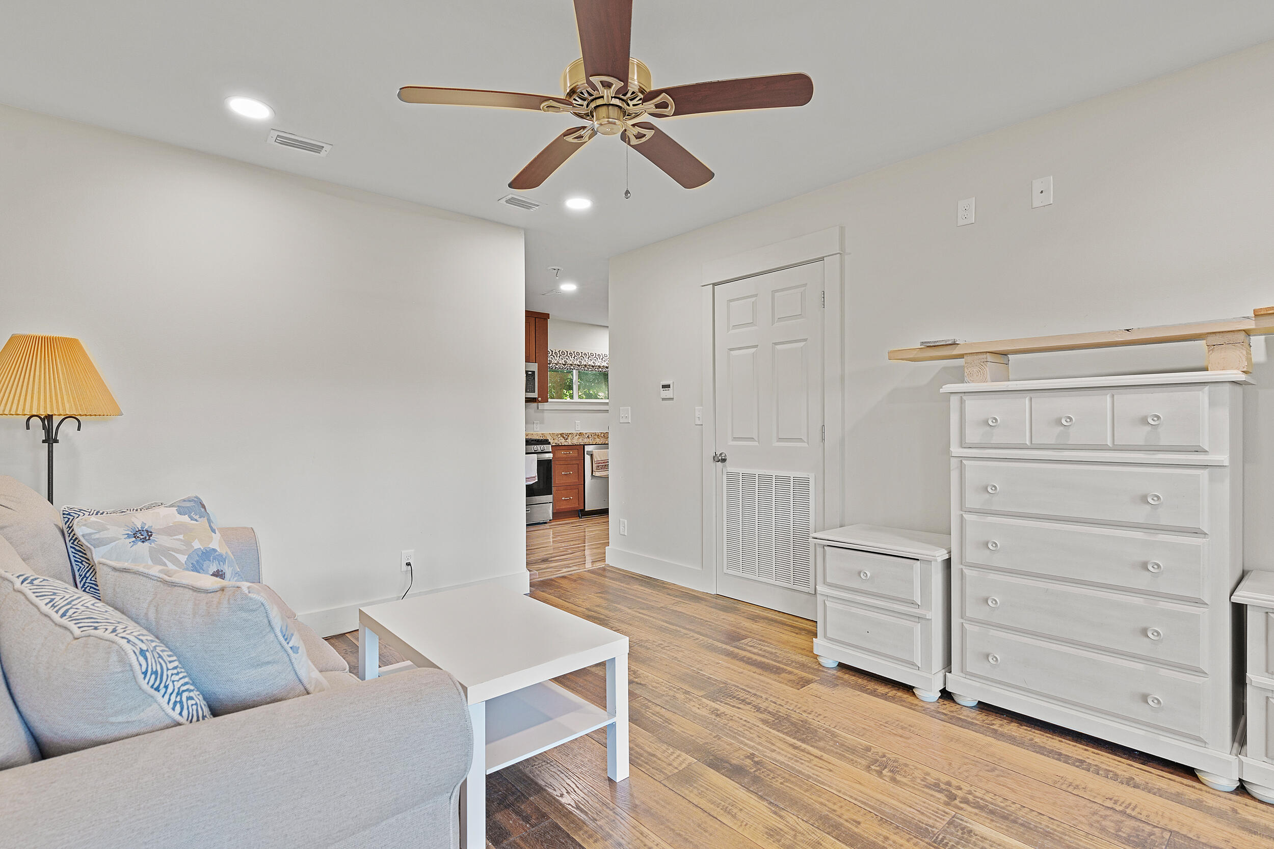 Furnished living area with a gray sofa, white dresser, wood-look floors, and a view into the kitchen.