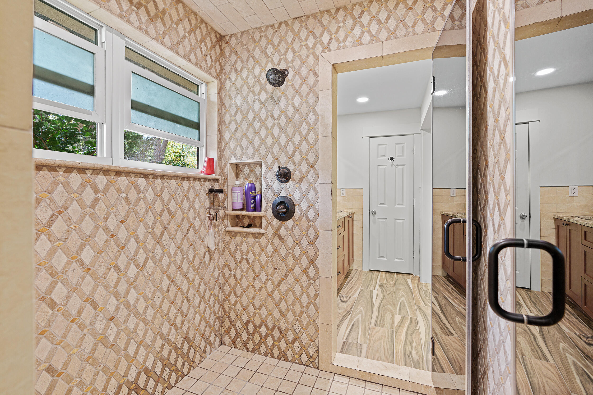 Walk-in shower with patterned stone tile, glass enclosure, dark fixtures, and two windows.