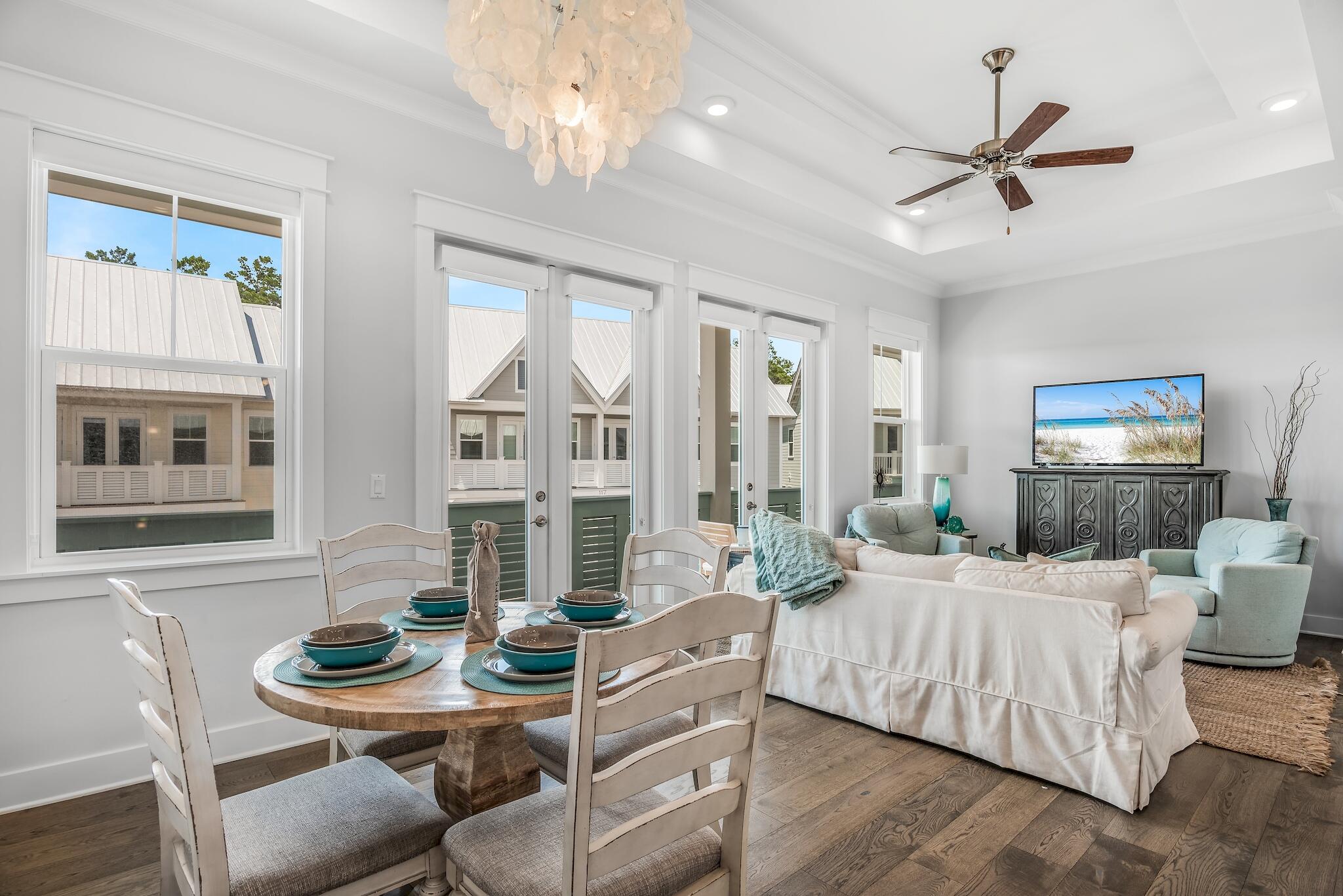 Open-plan living and dining room with a round set table, white sofa, pale blue chairs, and large windows.