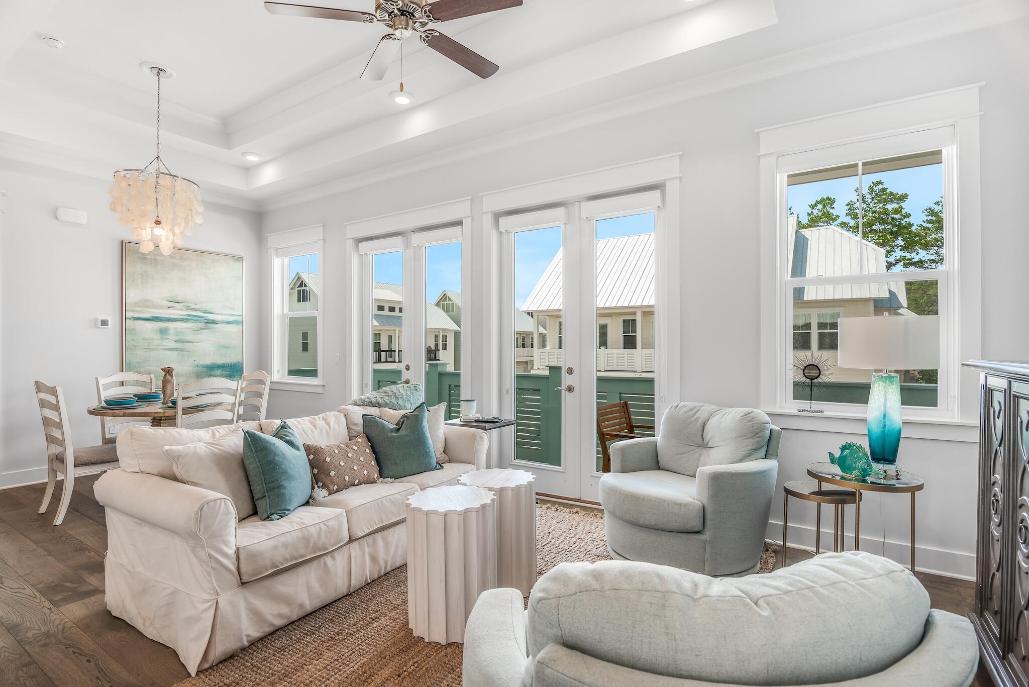 Furnished living room with white seating, teal pillows, and large windows overlooking nearby homes.