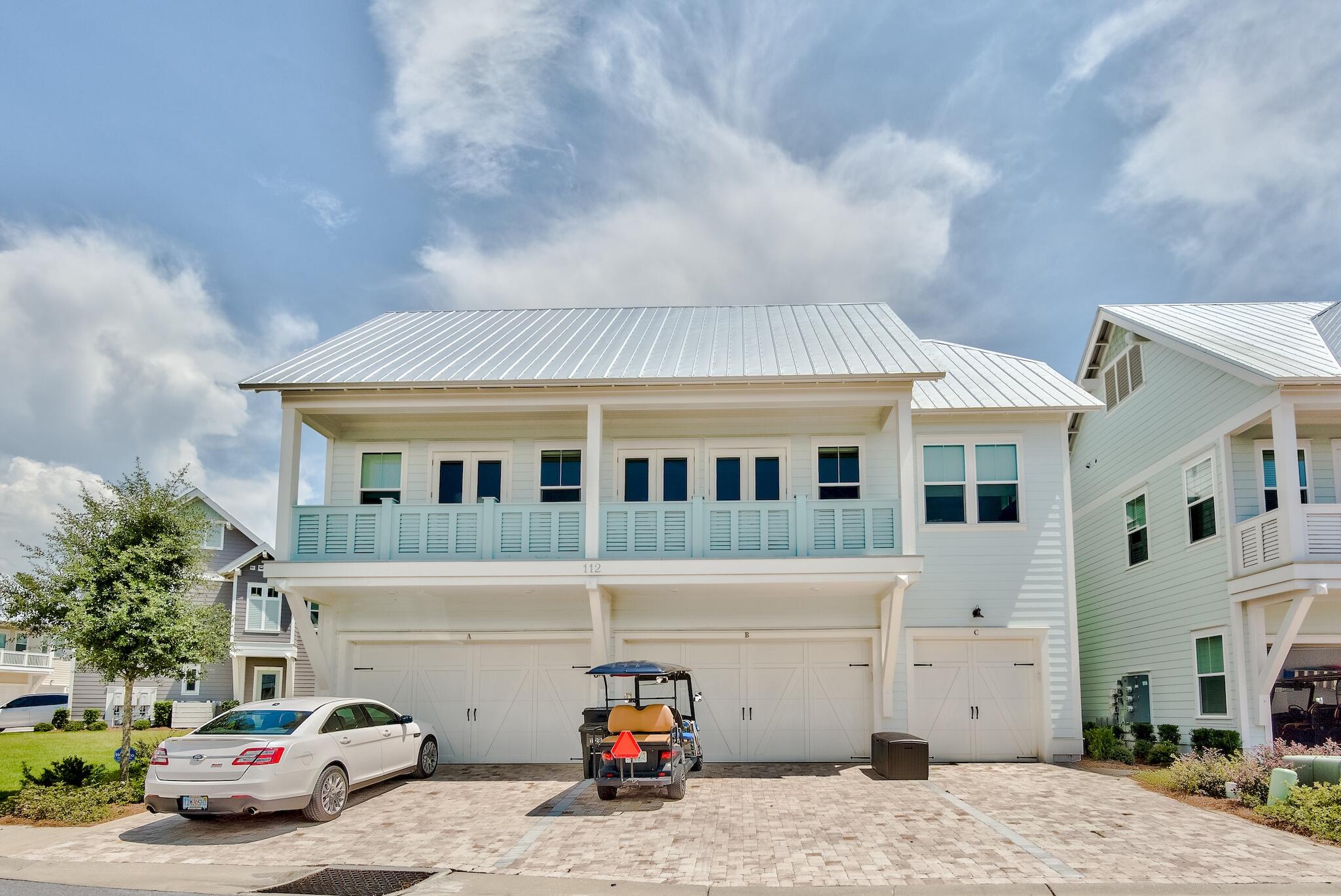 White multi-unit home with a metal roof, upper balconies, and three garage doors along a paved driveway.
