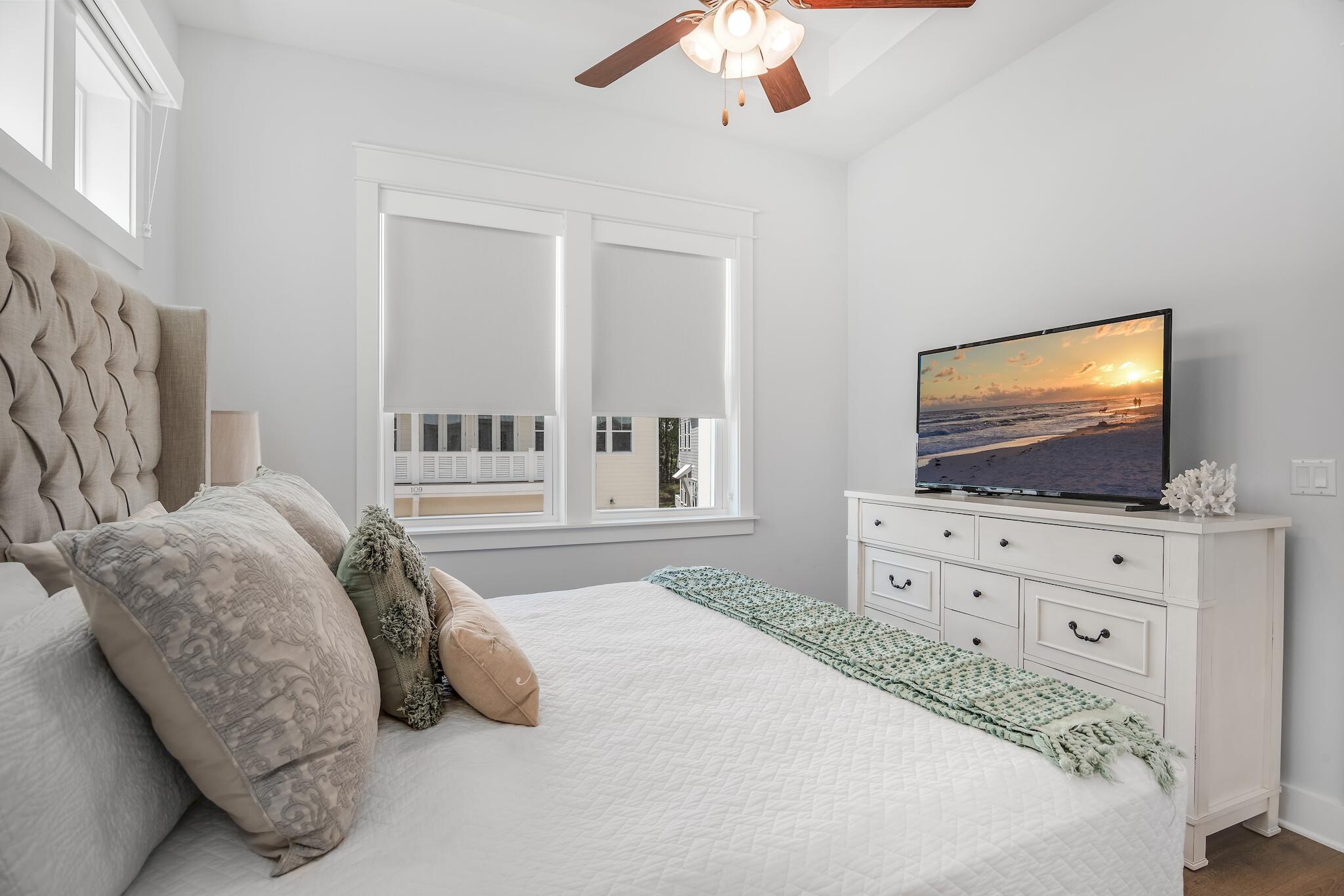 Furnished bedroom with a white quilted bed, tufted headboard, and television showing a beach sunset.