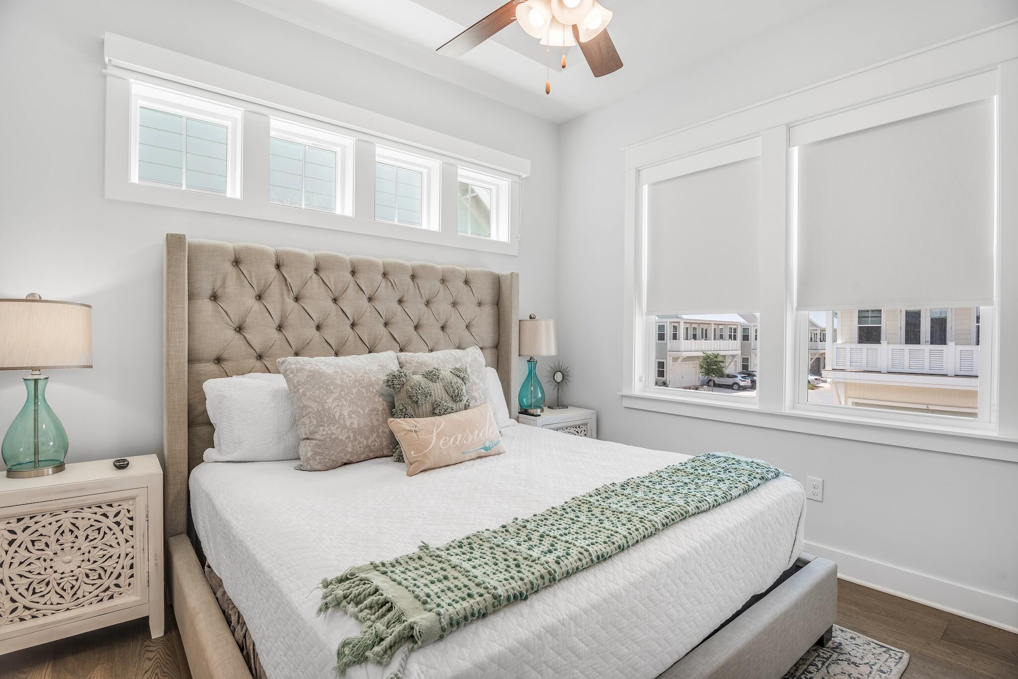 Staged bedroom with a tufted beige headboard, white bedding, green throw, and large windows overlooking nearby homes.