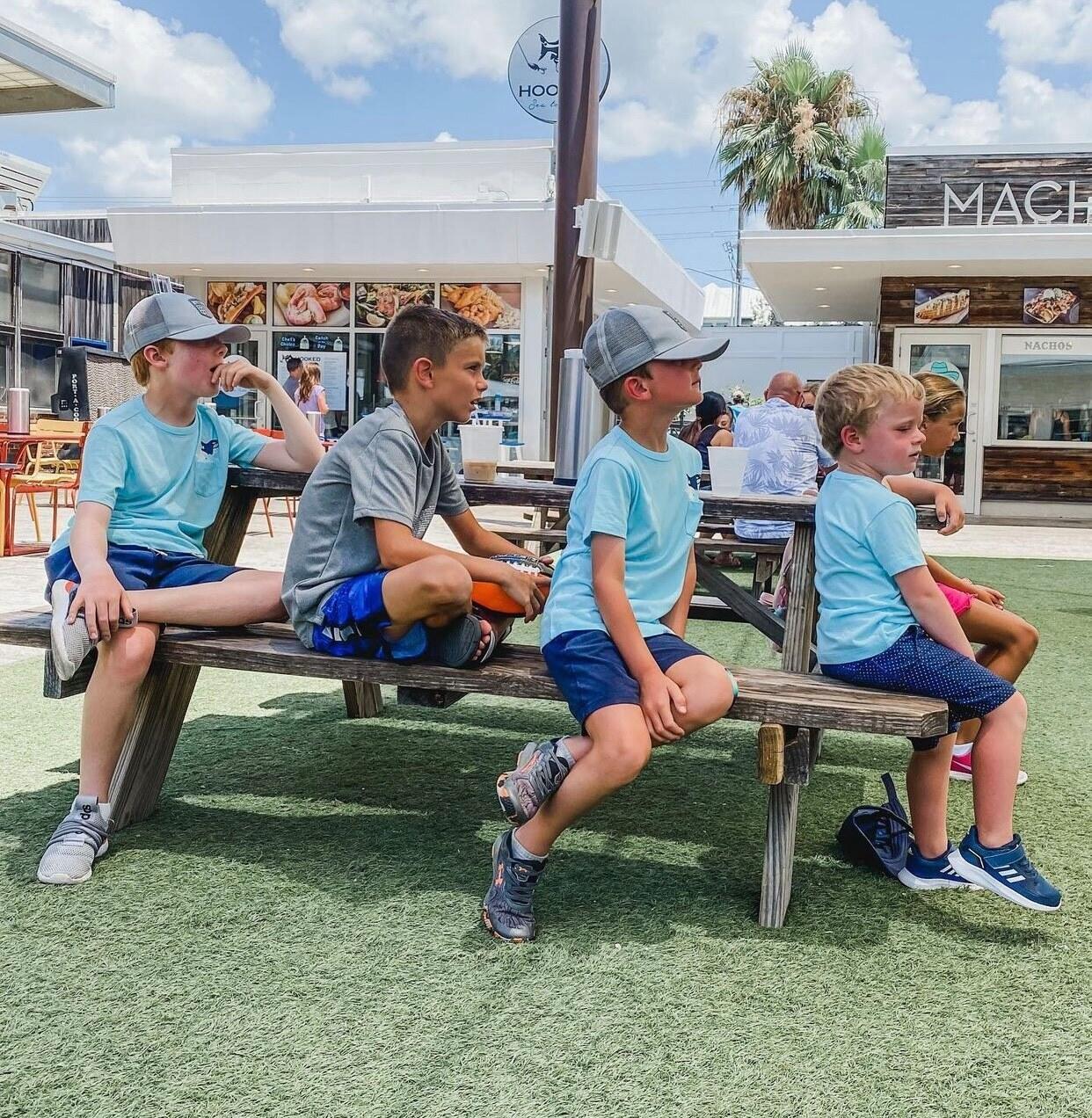 Four boys sit together on a picnic bench at an outdoor food court.