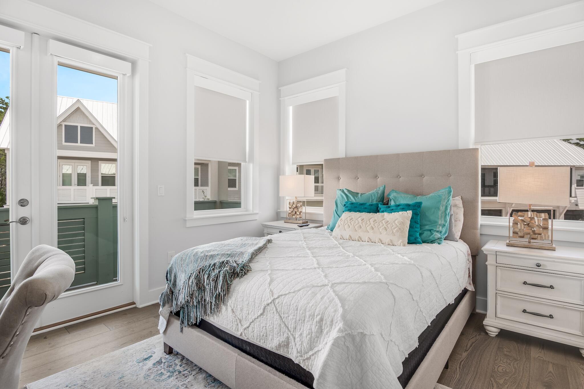 Bedroom with a white quilted bed, turquoise pillows, and windows overlooking neighboring homes.