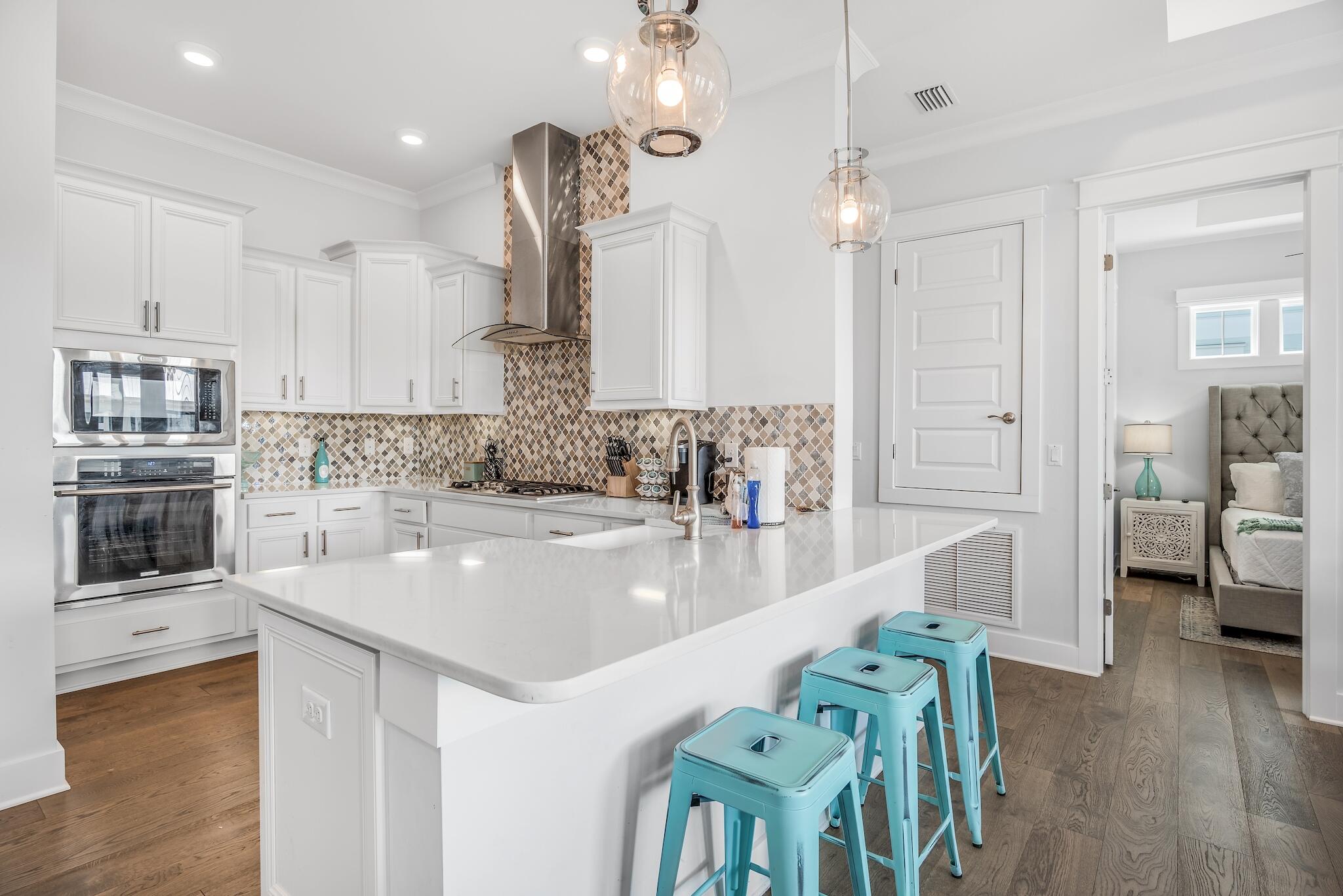 White kitchen with a large island, turquoise stools, patterned tile backsplash, and bedroom doorway.