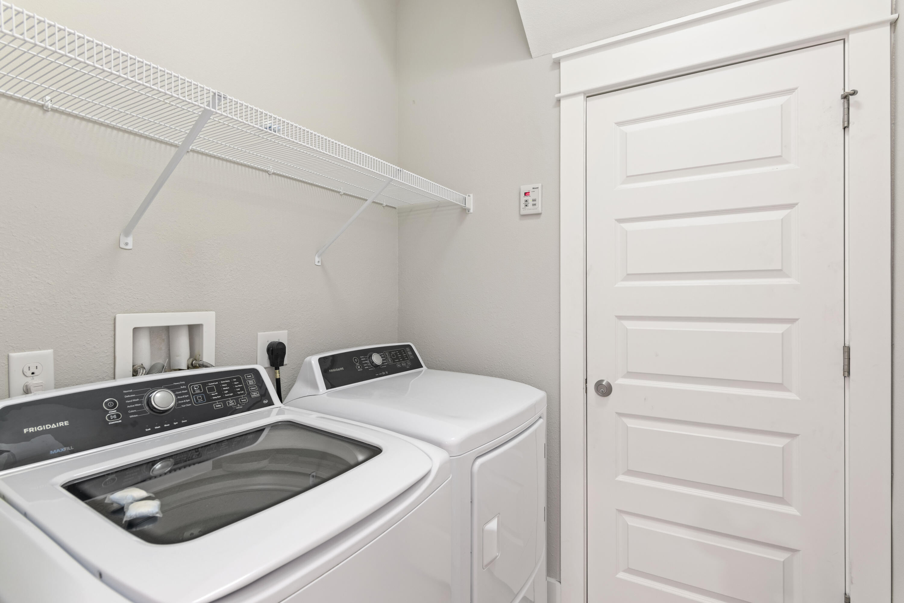 Laundry room with matching white washer and dryer beneath a wire shelf.