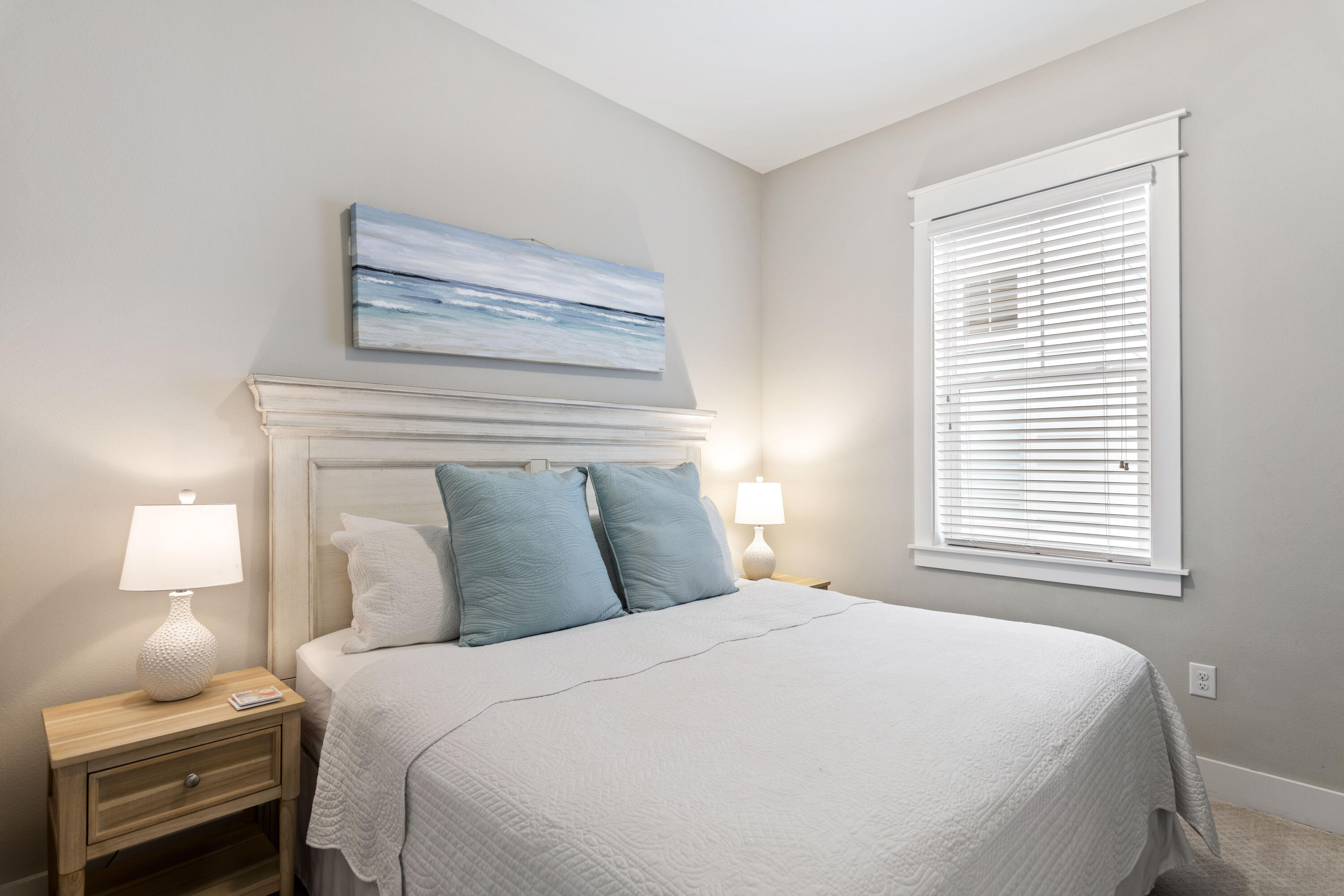 Bedroom with a white quilted bed, blue pillows, and beach painting above the headboard.