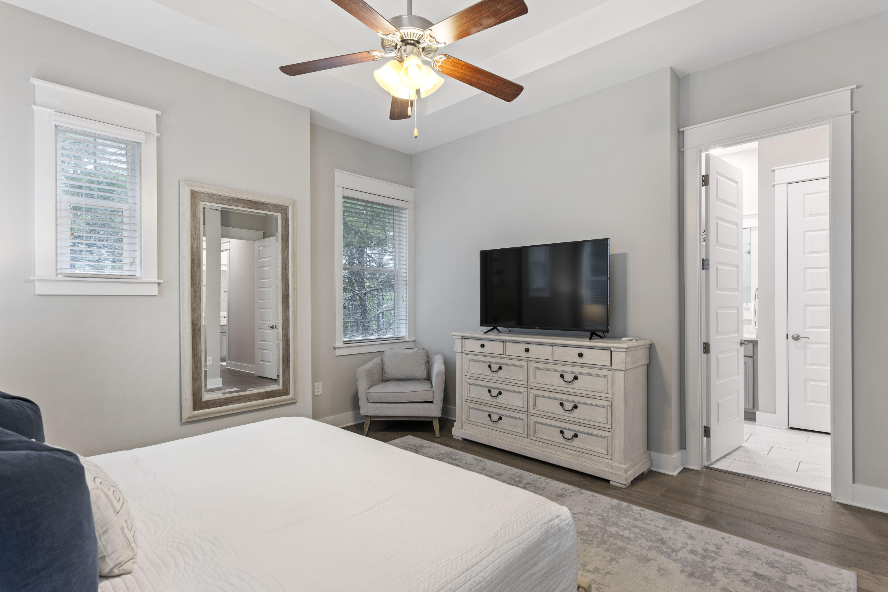 Furnished bedroom with a white bed, television dresser, ceiling fan, and adjoining bathroom doorway.