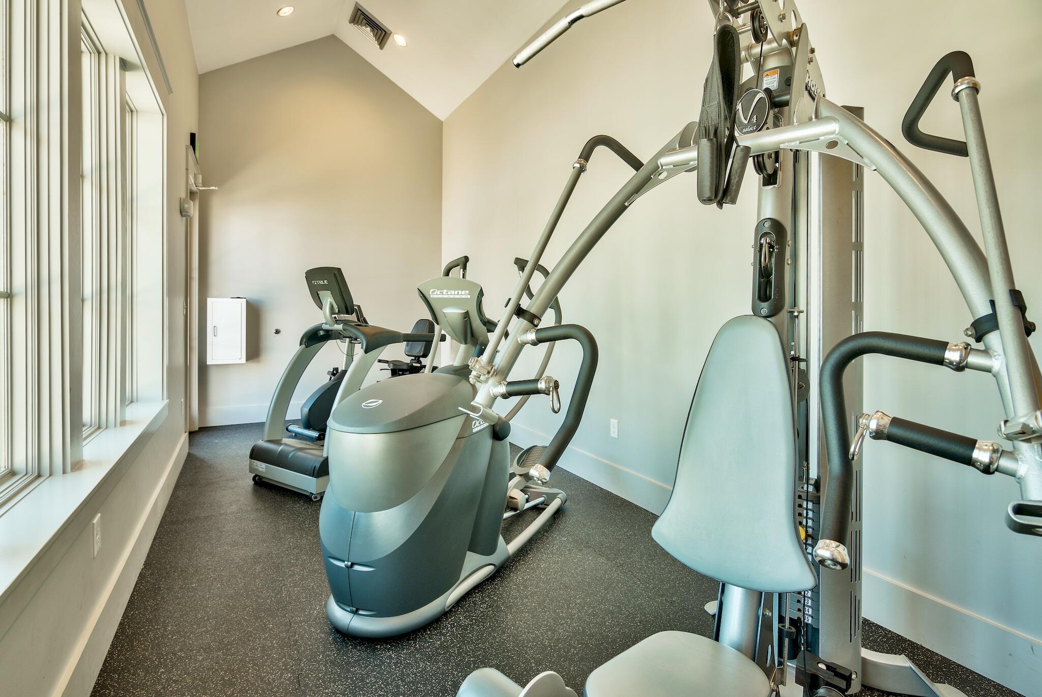 Fitness room with cardio machines and a cable weight-training machine beside large windows.