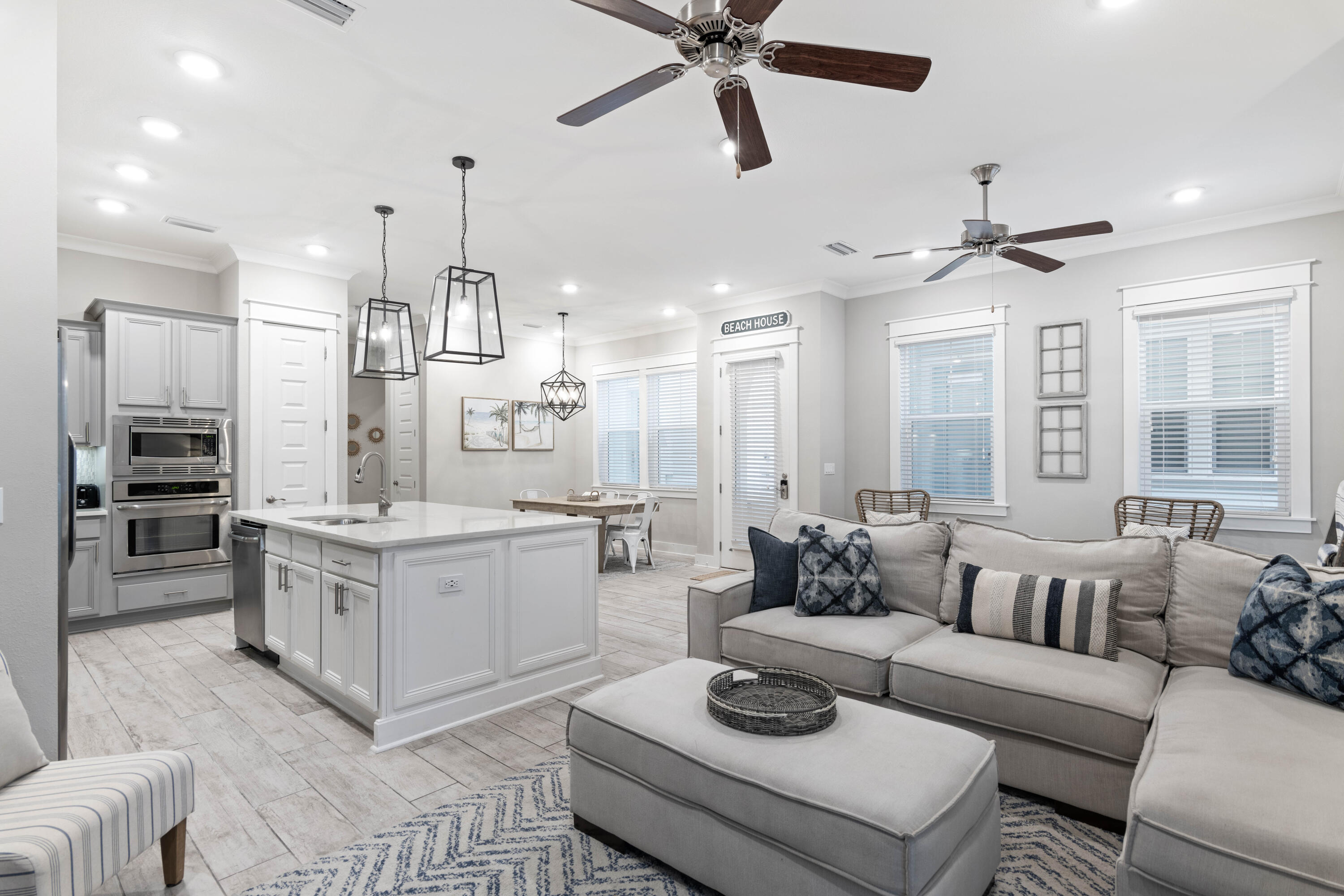 Open-plan living room and kitchen with a white island, gray sectional, and beach house sign.