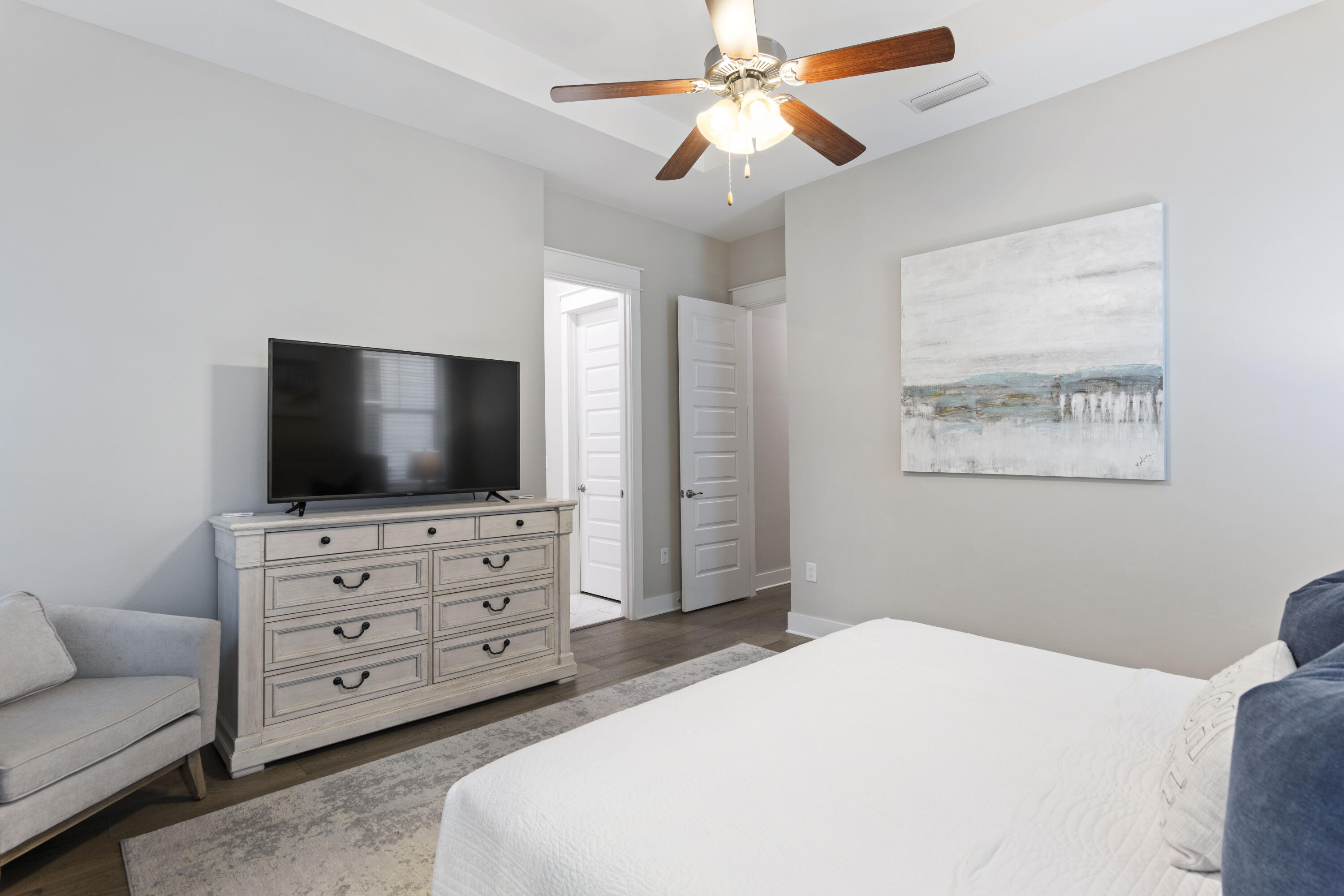 Furnished bedroom with a white bed, gray chair, and pale wood dresser topped by a television.