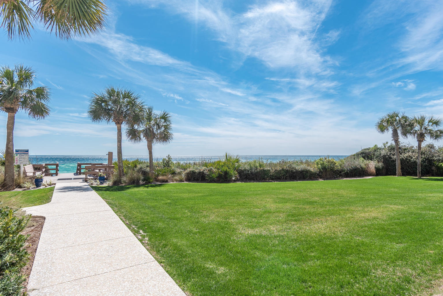 Landscaped beachfront lawn with a paved walkway leading to the ocean between palm trees.