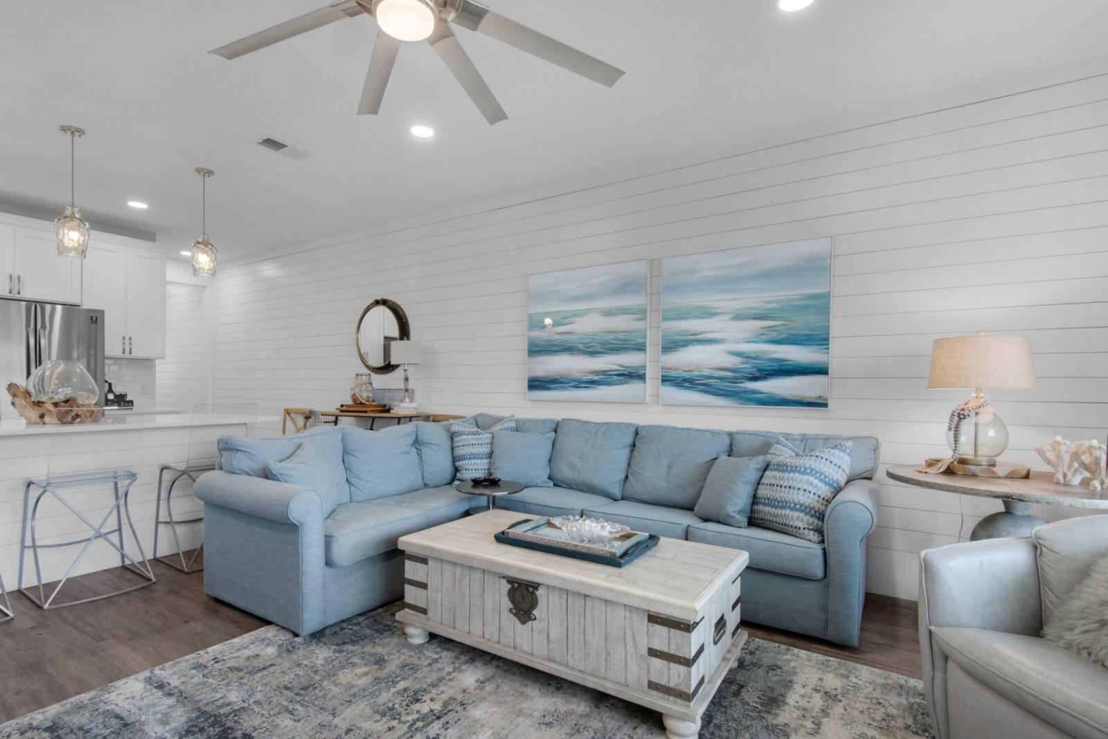 Living room with a pale blue sectional, coastal artwork, and white plank walls.