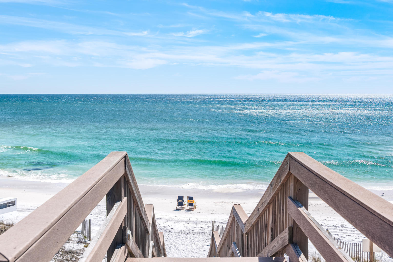Beach access stairs lead to white sand, two beach chairs, and turquoise ocean water.