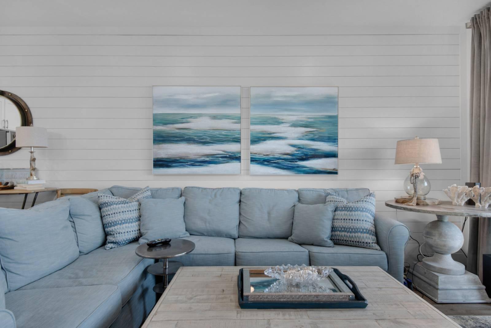 Living room with a pale blue sectional sofa beneath two ocean paintings on a white shiplap wall.