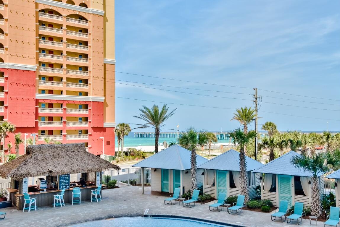 Beachfront condominium tower overlooking cabanas, a pool deck, turquoise water, and a distant pier.
