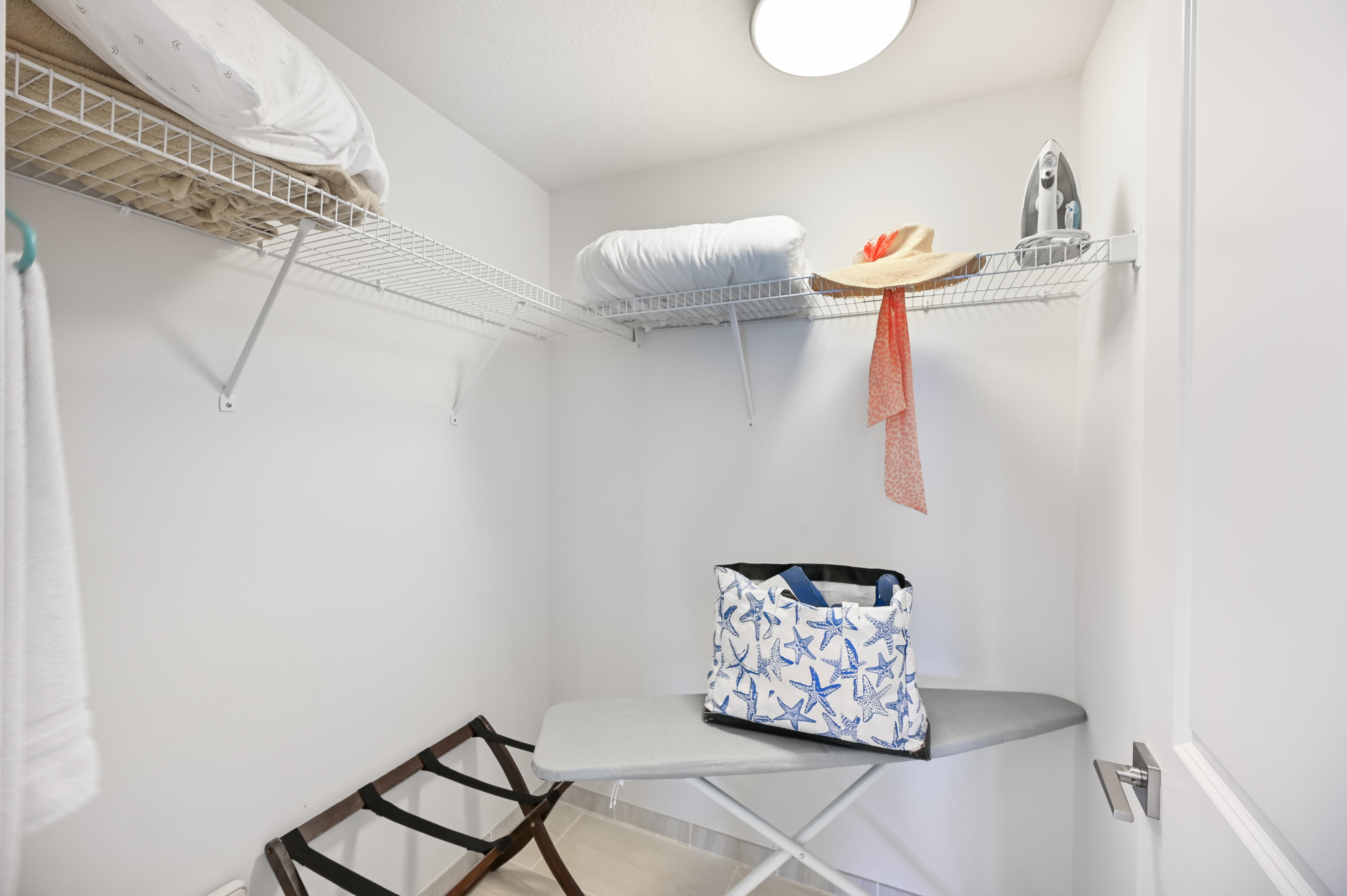 Walk-in closet with wire shelves, folded linens, an iron, and a starfish-patterned beach bag on an ironing board.