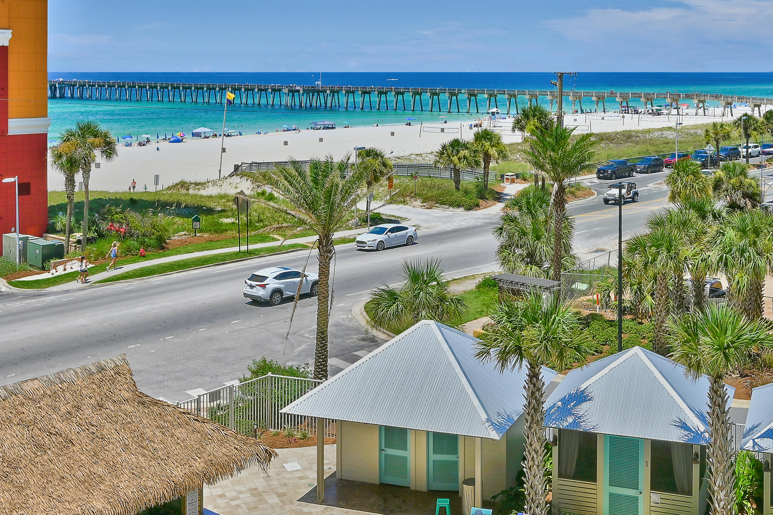 Panoramic beach view with turquoise water, a long pier, palm trees, and a coastal road.