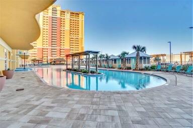 Curved condominium pool beside a tiled deck, lounge chairs, palm trees, and a tall beachfront tower.