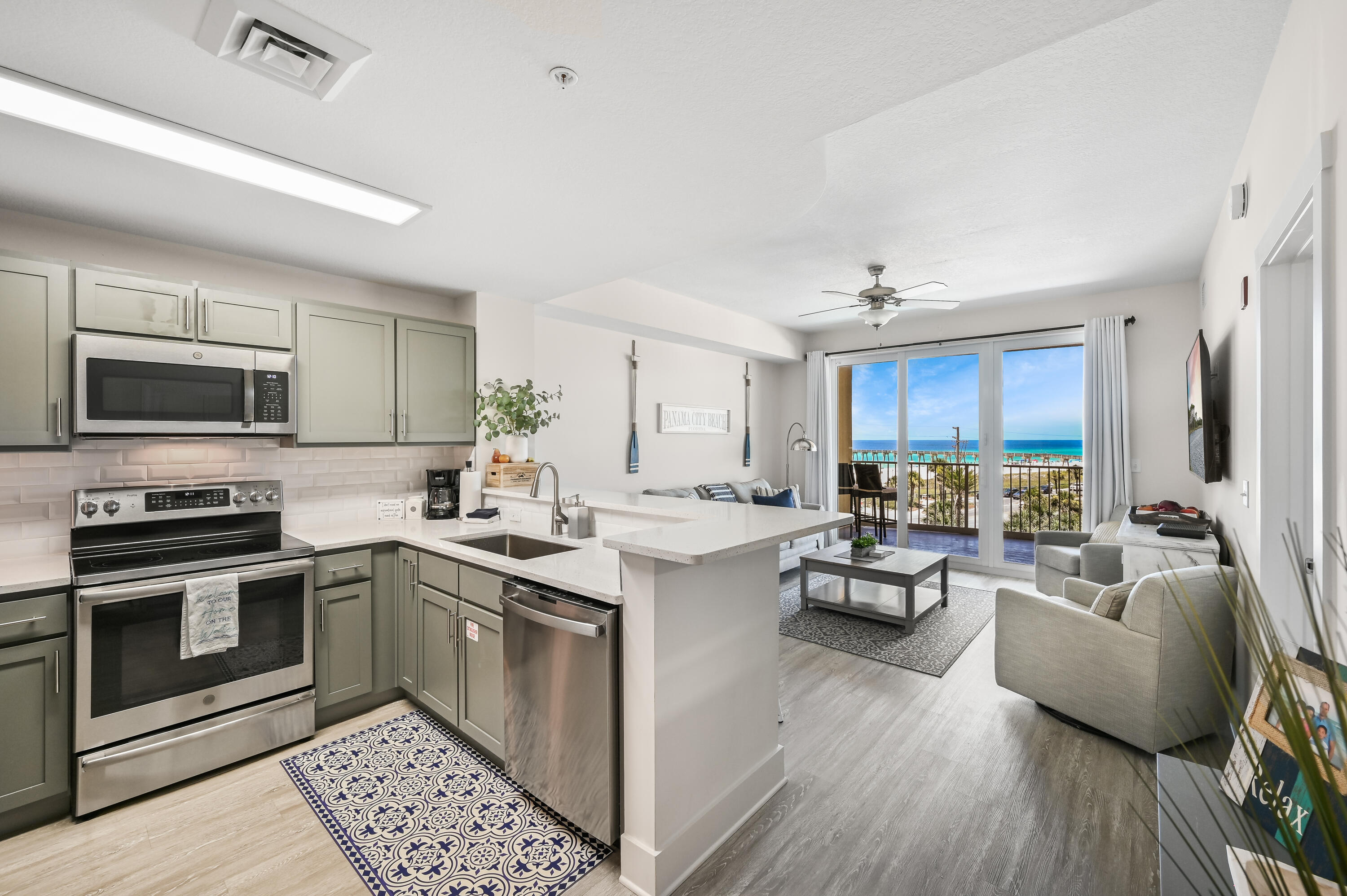 Open-plan condo kitchen and living room with sage-green cabinets and an ocean view from the balcony.