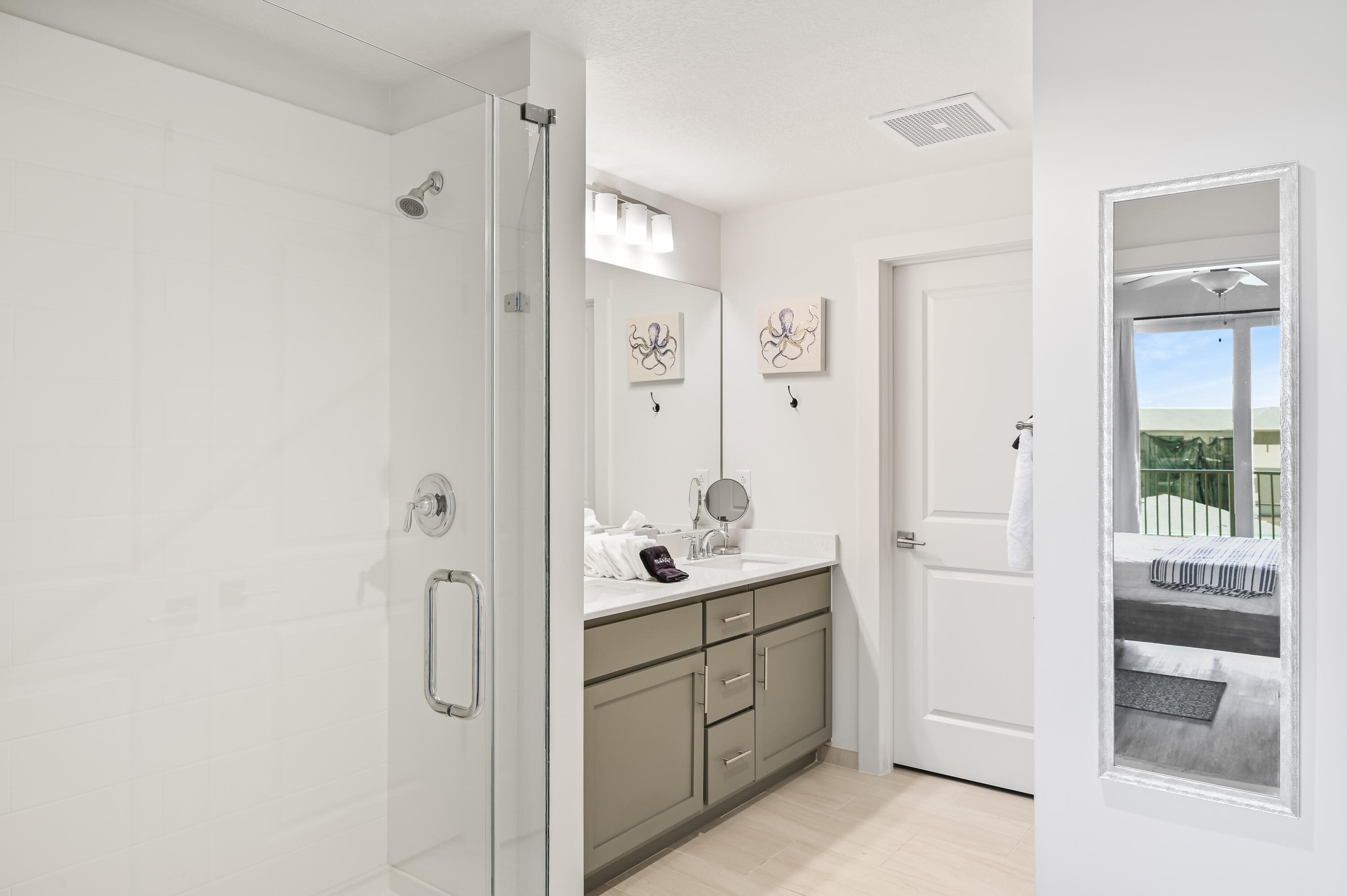Bathroom with glass shower and gray vanity; mirror reflects a bedroom and balcony.
