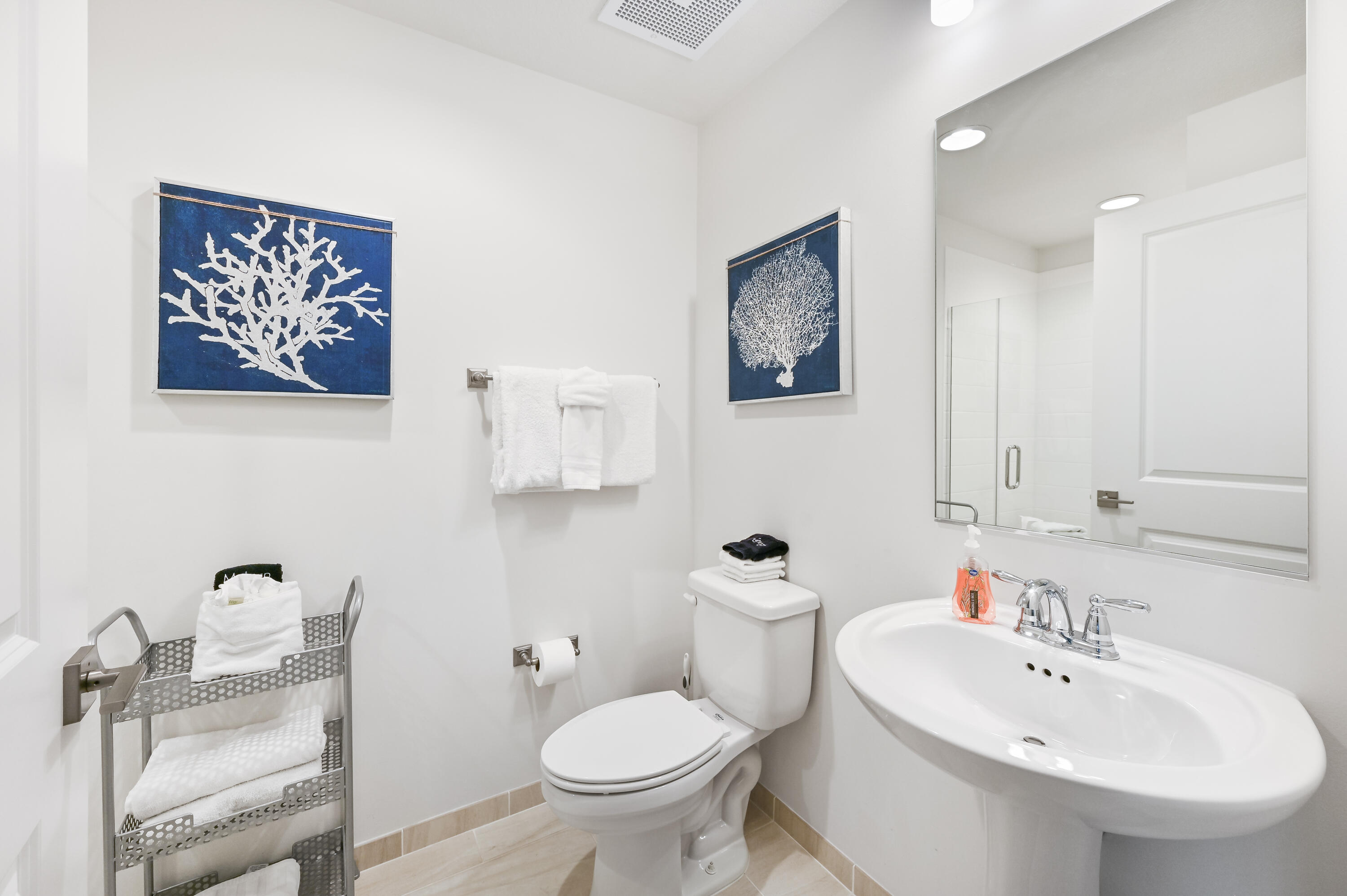 Bright bathroom with a pedestal sink, toilet, blue coral artwork, and neatly arranged white towels.