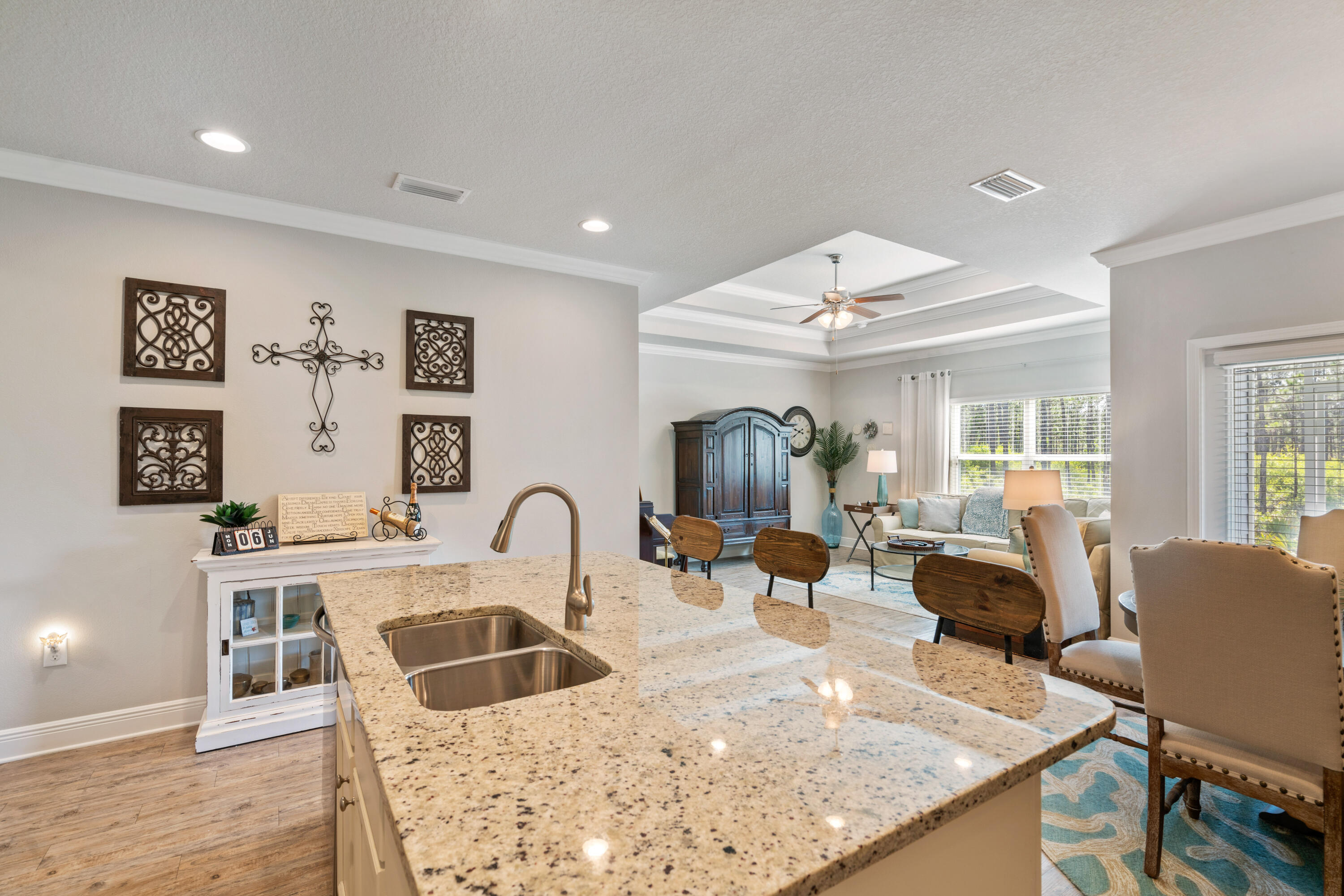 Granite kitchen island with double sink overlooks a furnished living room with a tray ceiling and large windows.
