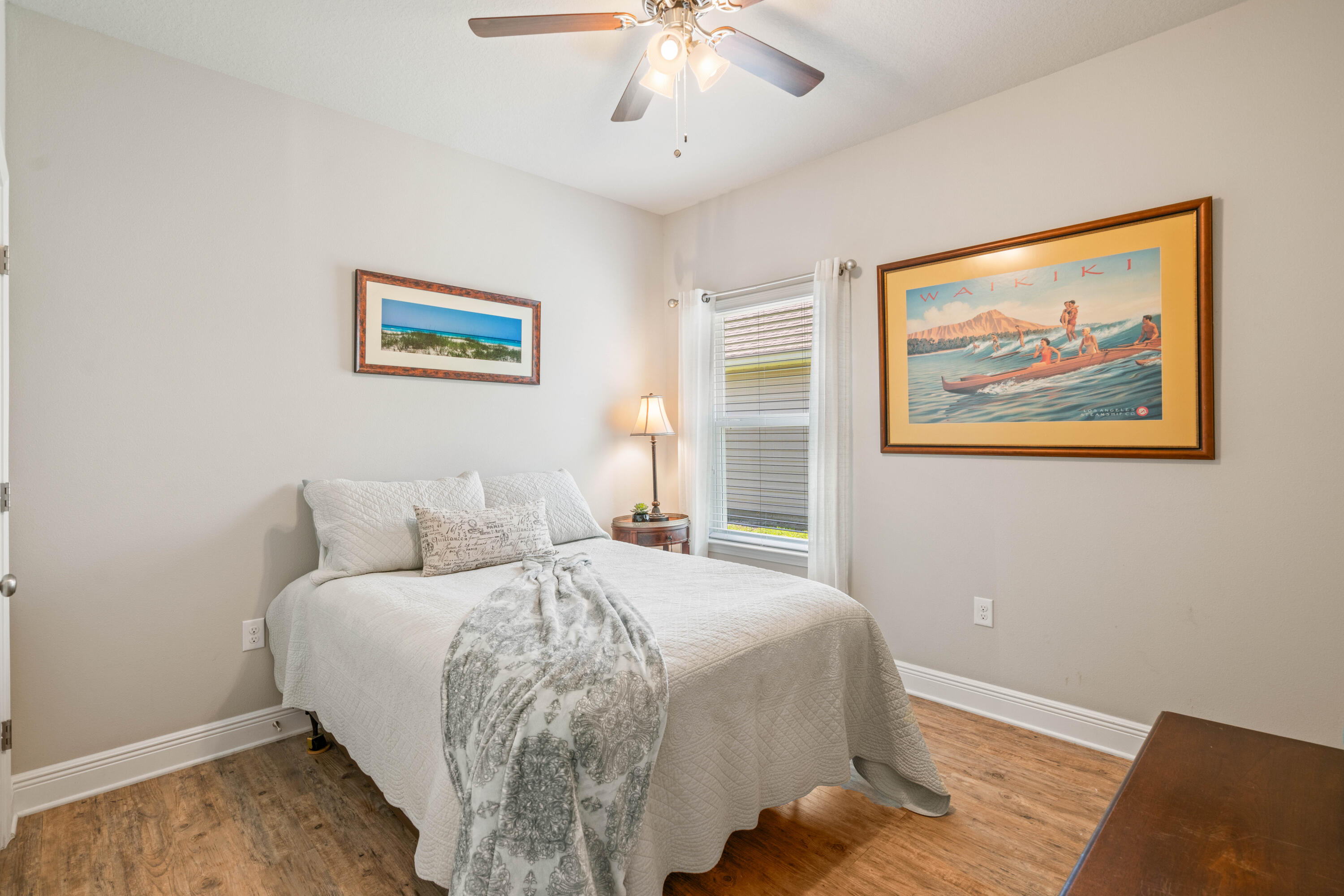 Bedroom with a white quilted bed, wood-look flooring, and framed Waikiki surfing artwork.