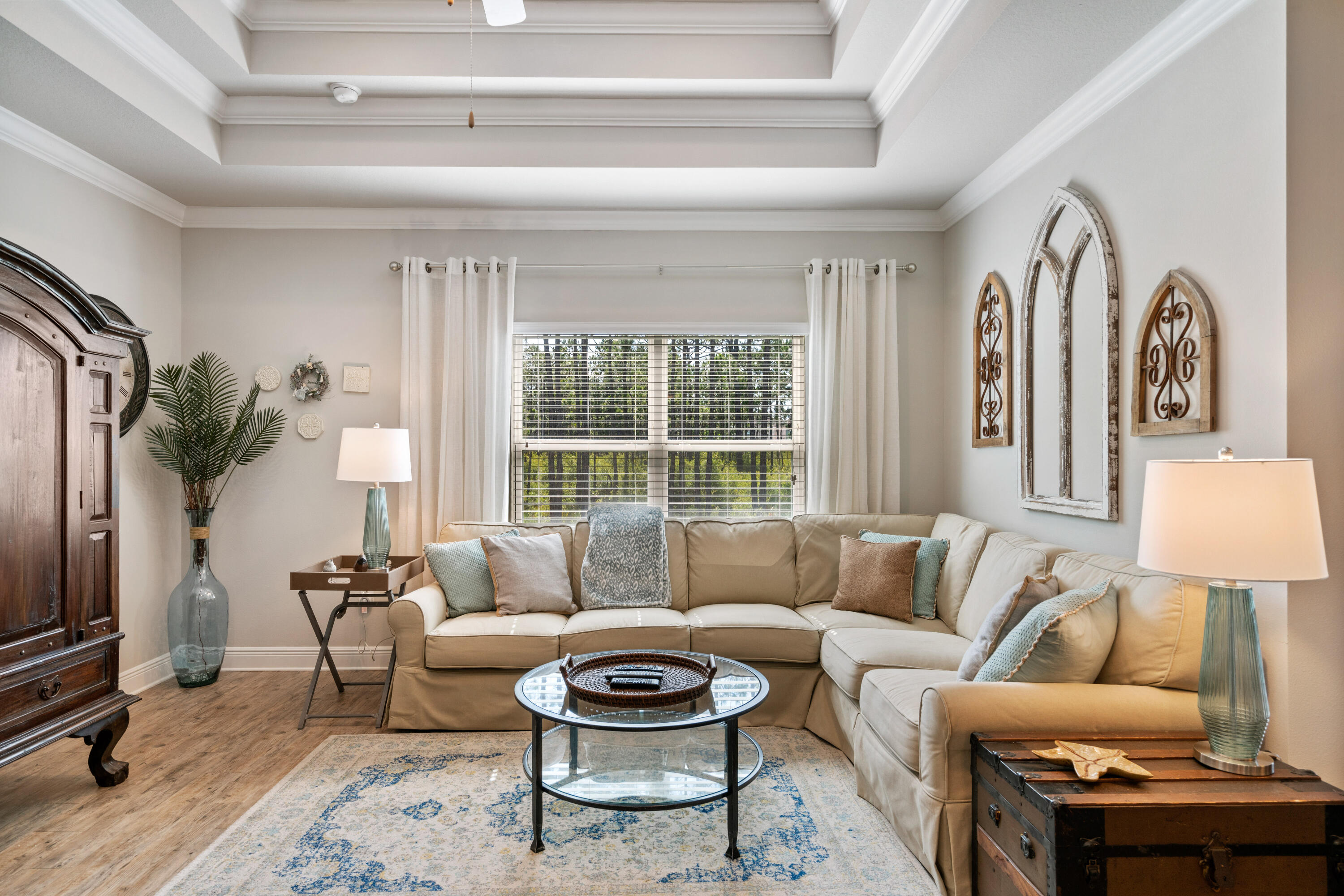 Living room with a beige sectional, glass coffee table, patterned rug, and large window overlooking greenery.
