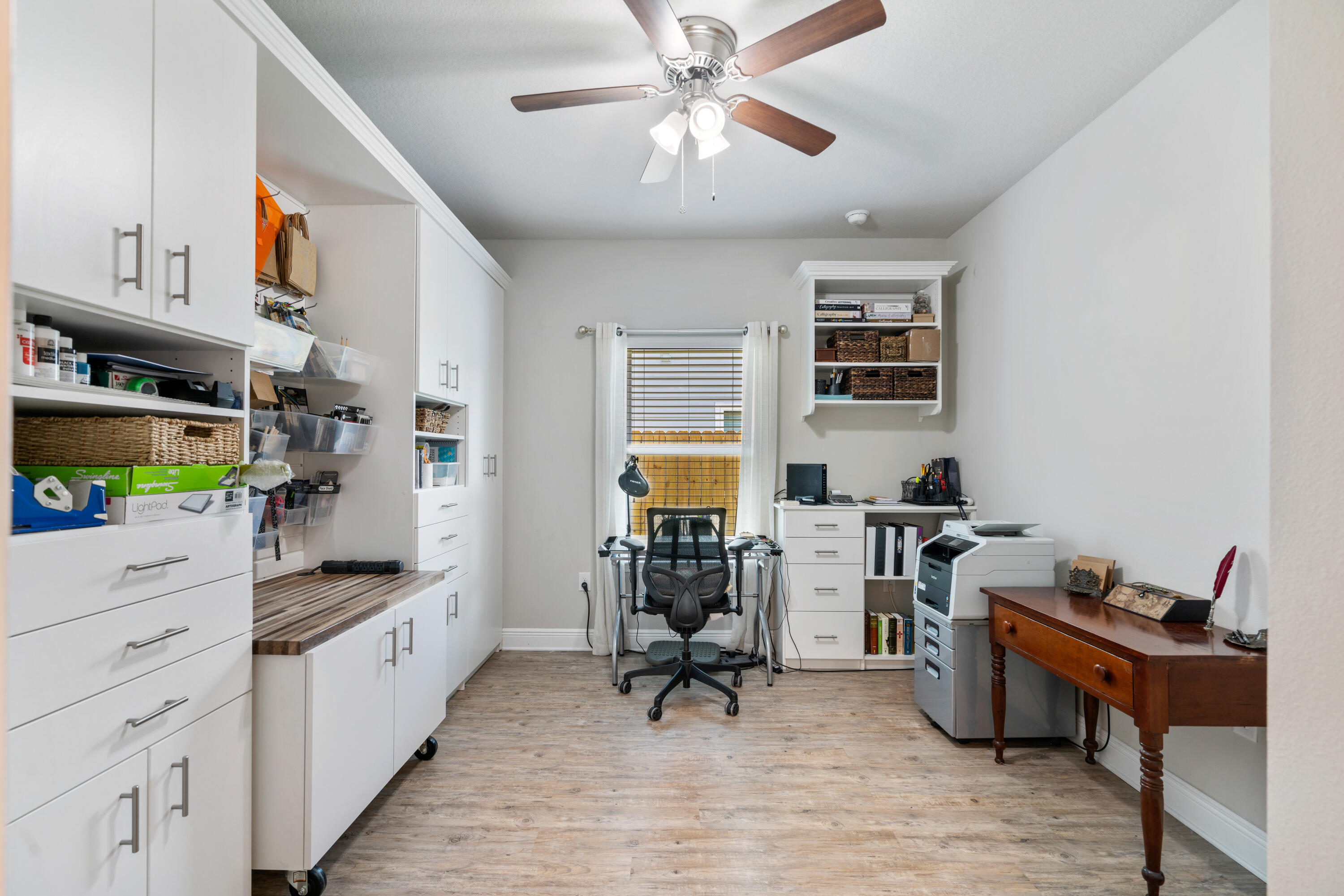 Home office with white built-in storage, a desk beneath the window, and a printer beside a wooden writing desk.